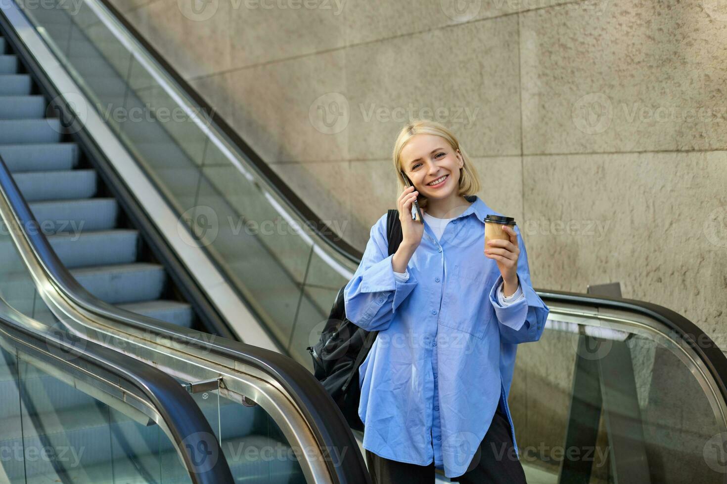 Smiling young woman on escalator, talking on mobile phone, drinking coffee and looking happy, going to university or college, on her way in city photo