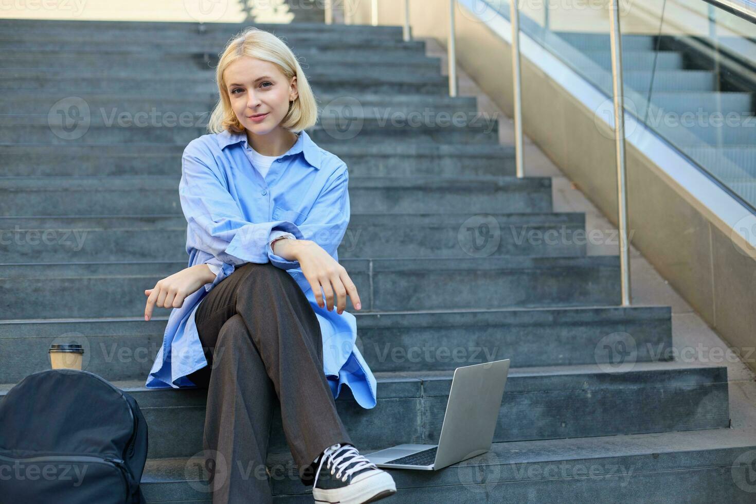 Portrait of young urban woman, college student with backpack, laptop and coffee, sitting on stairs in city, resting outdoors, taking break photo