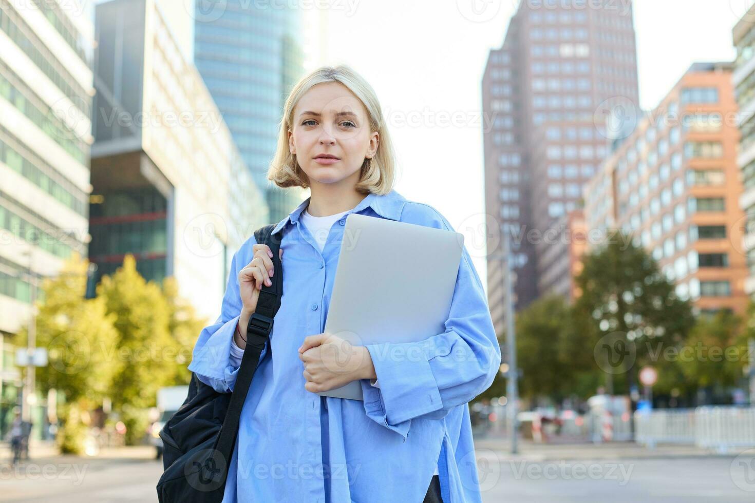 retrato de joven confidente mujer, Universidad estudiante con mochila y computadora portátil, Bóveda a lección, en pie al aire libre en vacío calle con grande edificios detrás foto