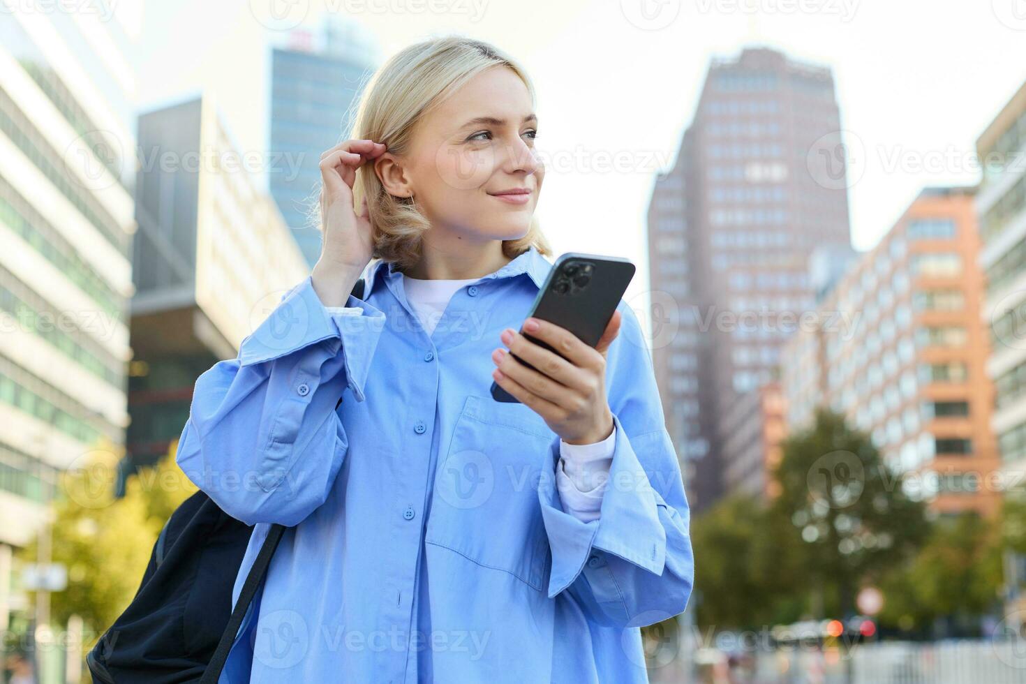 Portrait of beautiful young woman in city centre, standing in blue shirt with smartphone, checking notifications, waiting for car, order taxi on mobile phone app photo