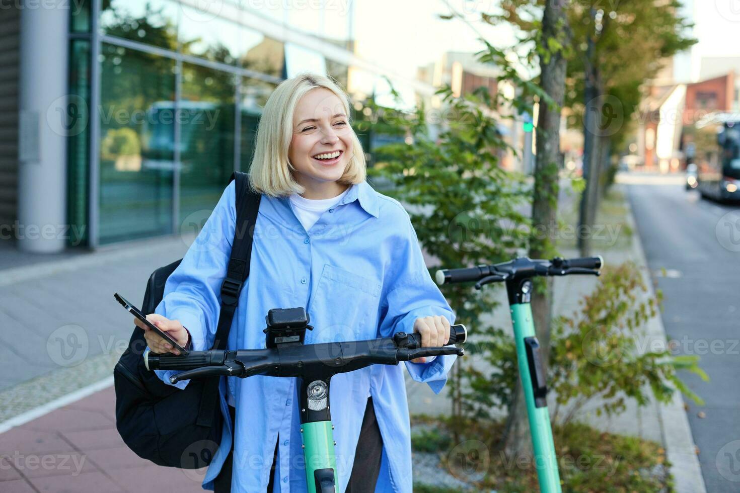 Portrait of smiling blond woman, college student using smartphone app to scan QR code on green electric scooter on street, renting a ride photo