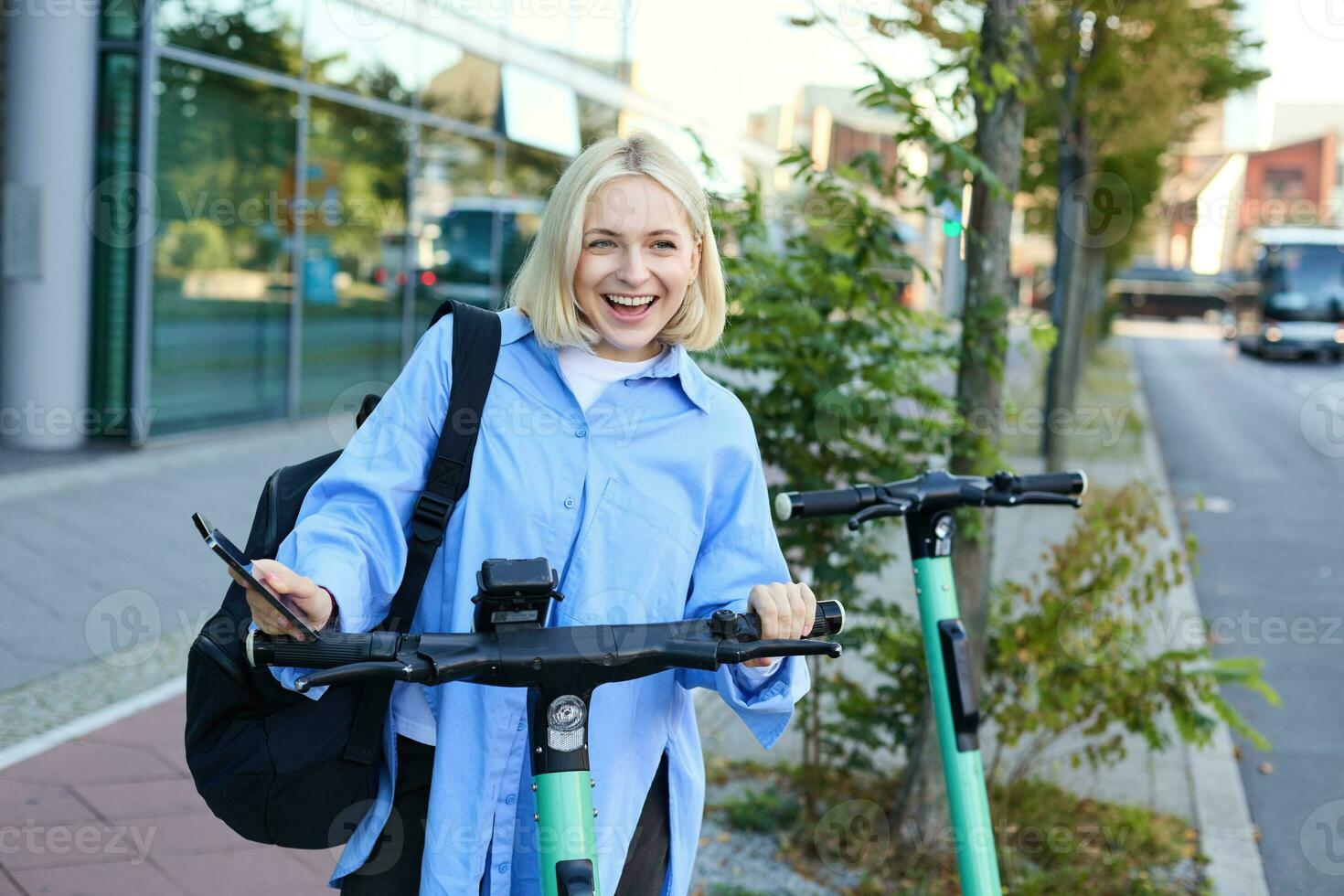 Portrait of young smiling woman, unlocks electric scooter, scans qr code with smartphone app to unlock her ride, standing on street near building photo