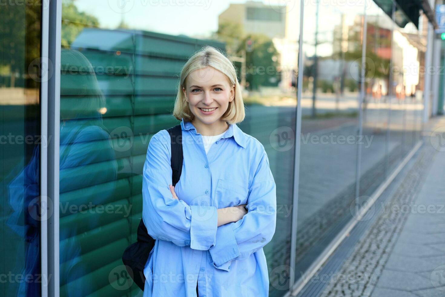 Image of young modern woman on street, college student in blue shirt, with backpack, cross arms on chest and smiles with confidence at camera photo