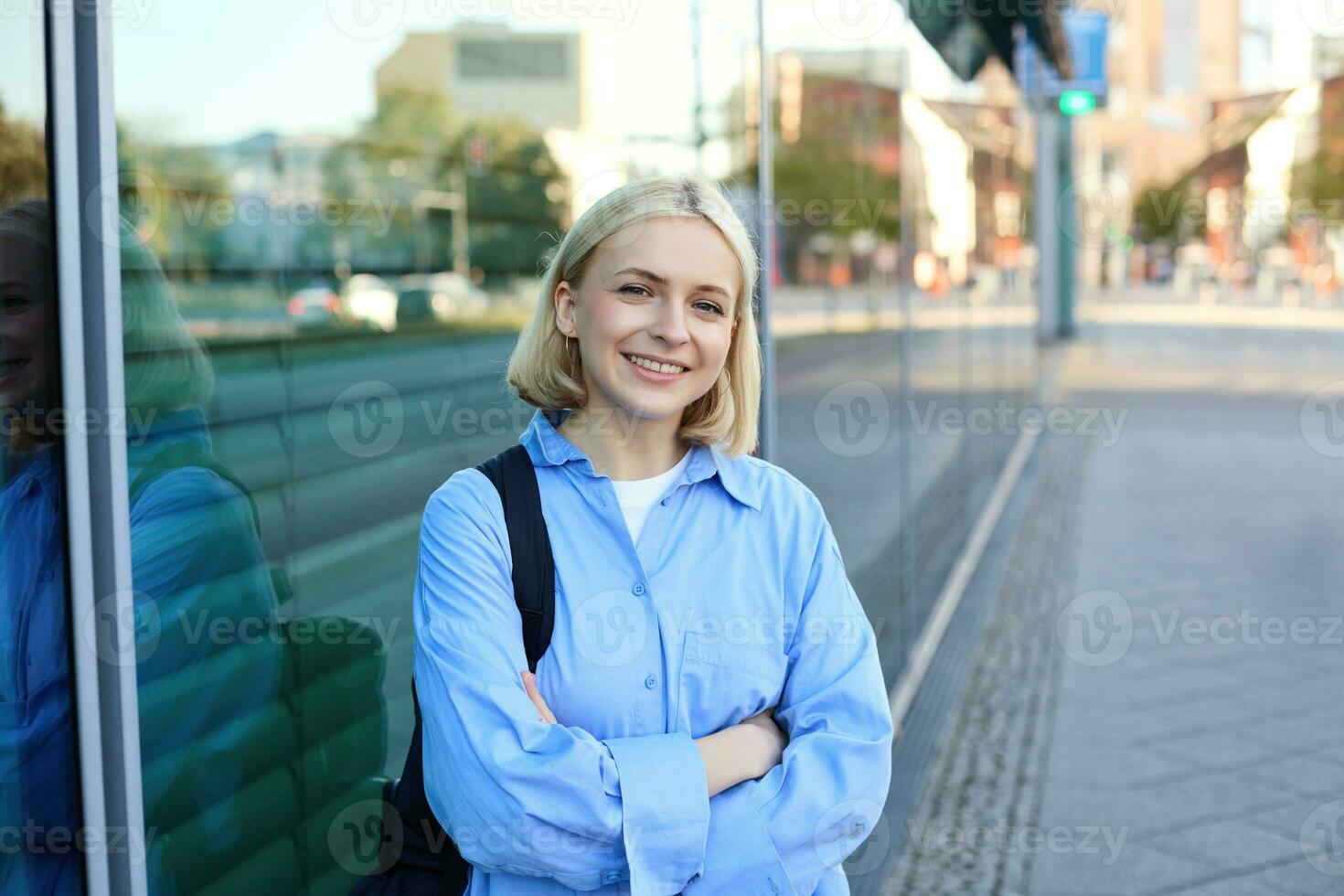 Lifestyle portrait of young smiling woman, student or office employee, standing on street in blue shirt, cross arms on chest and looking confident at camera photo