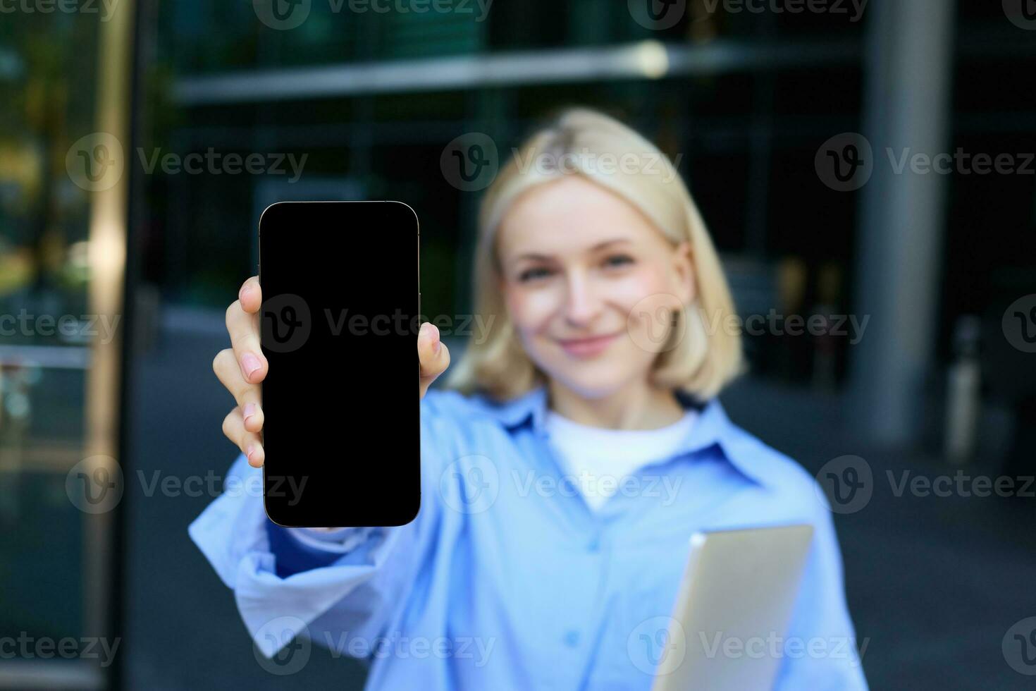 retrato de hermosa joven mujer, estudiante posando en calle cerca instalaciones, participación computadora portátil, demostración móvil teléfono pantalla, demostrando aplicación o promoción en teléfono inteligente y sonriente foto