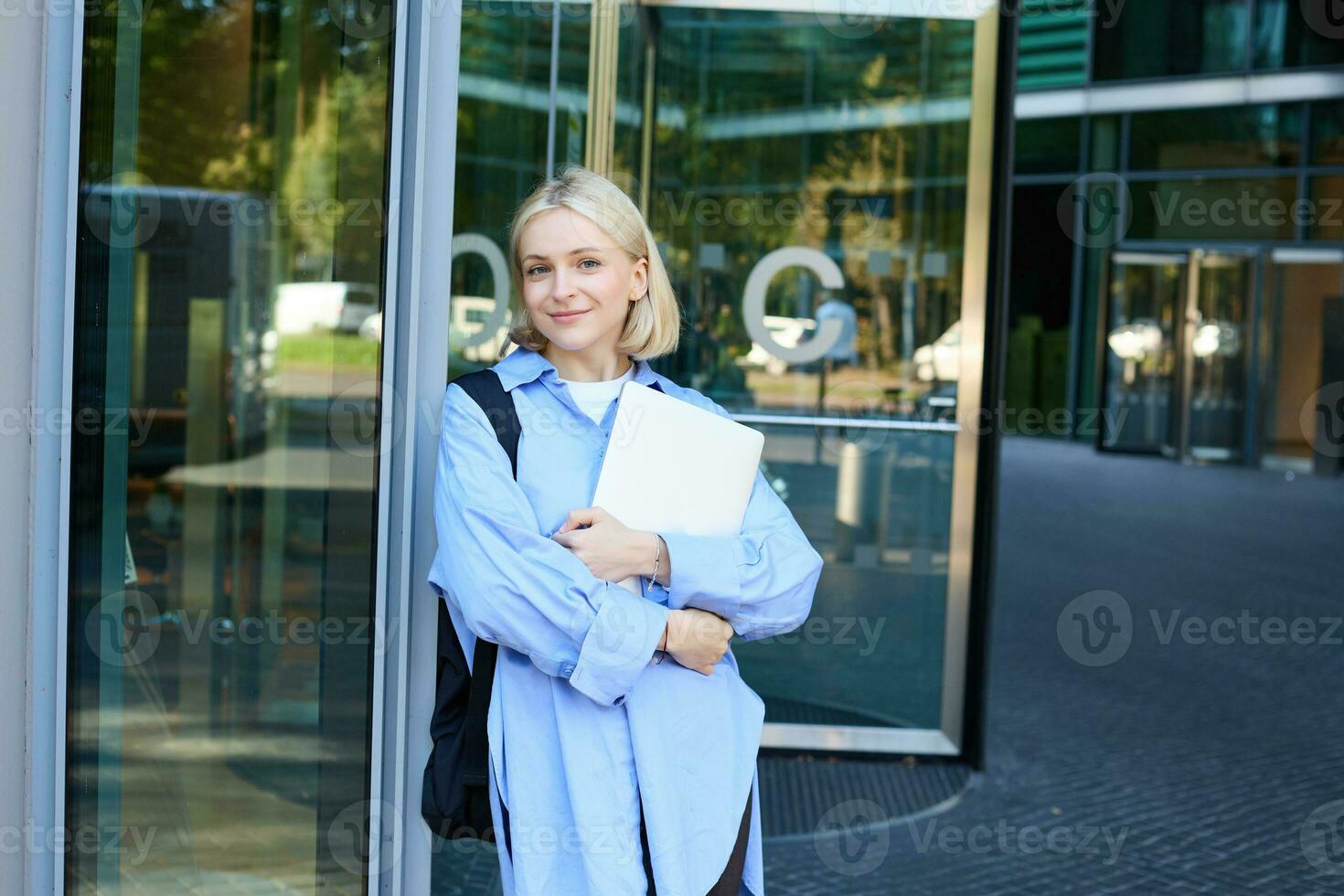 Lifestyle portrait of young woman with laptop, posing near campus, entrance to office building, holding backpack, smiling and looking happy photo