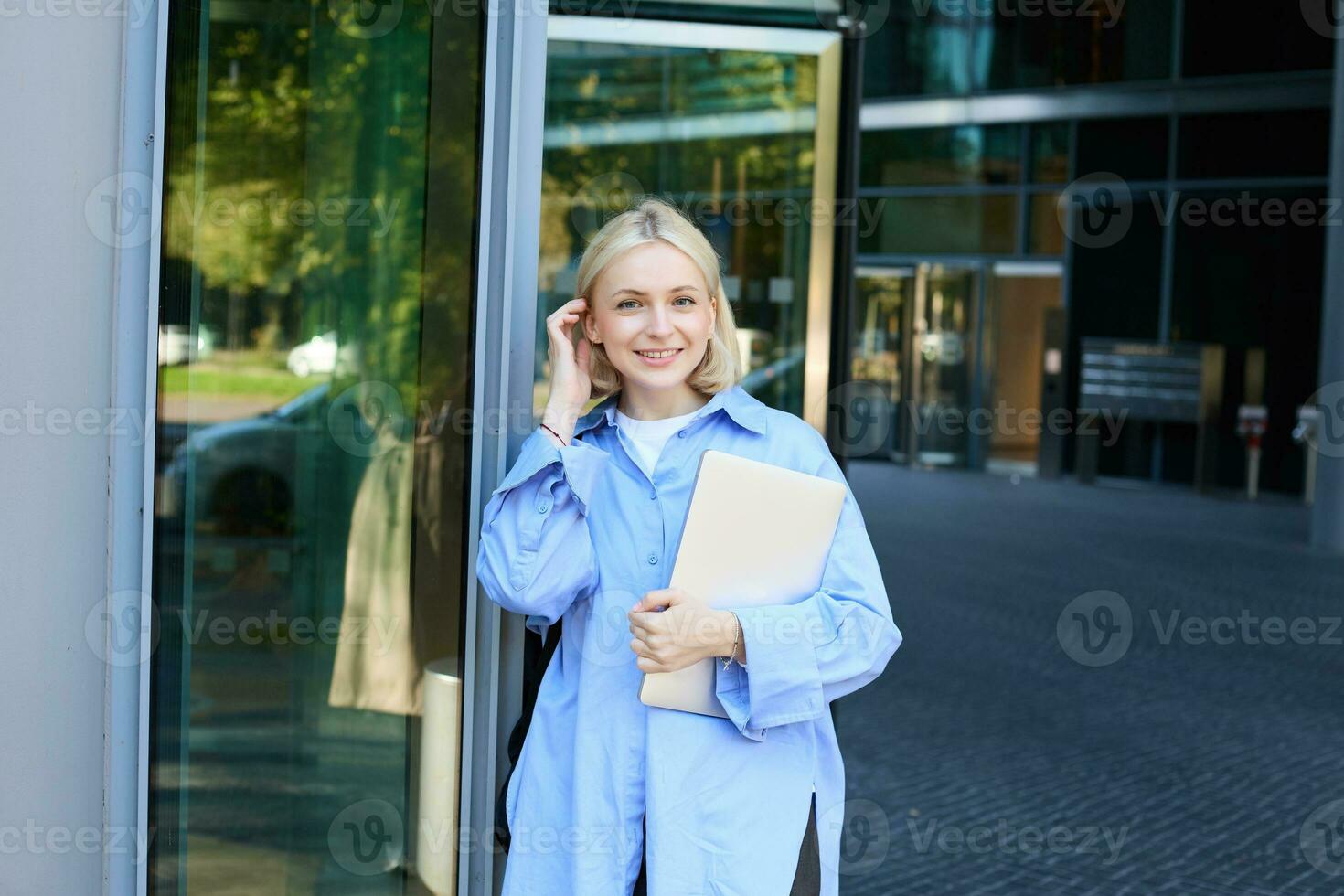 Lifestyle portrait of young woman with laptop, posing near campus, entrance to office building, holding backpack, smiling and looking happy photo