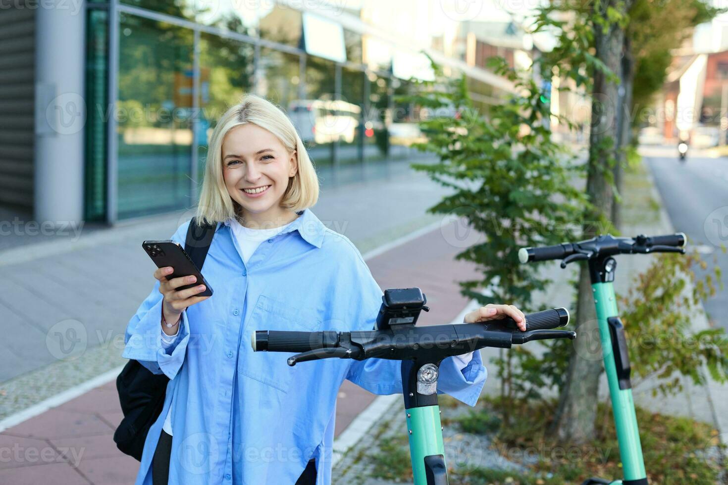 Portrait of young woman, renting a scooter, using mobile phone app to unlock it, using quick ride to get to work, smiling and looking happy photo