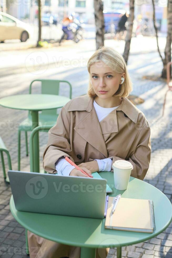 Vertical shot of young blond woman in wireless headphones, making notes, studying online, attend web lecture, using laptop, writing in notebook, looking serious at camera photo