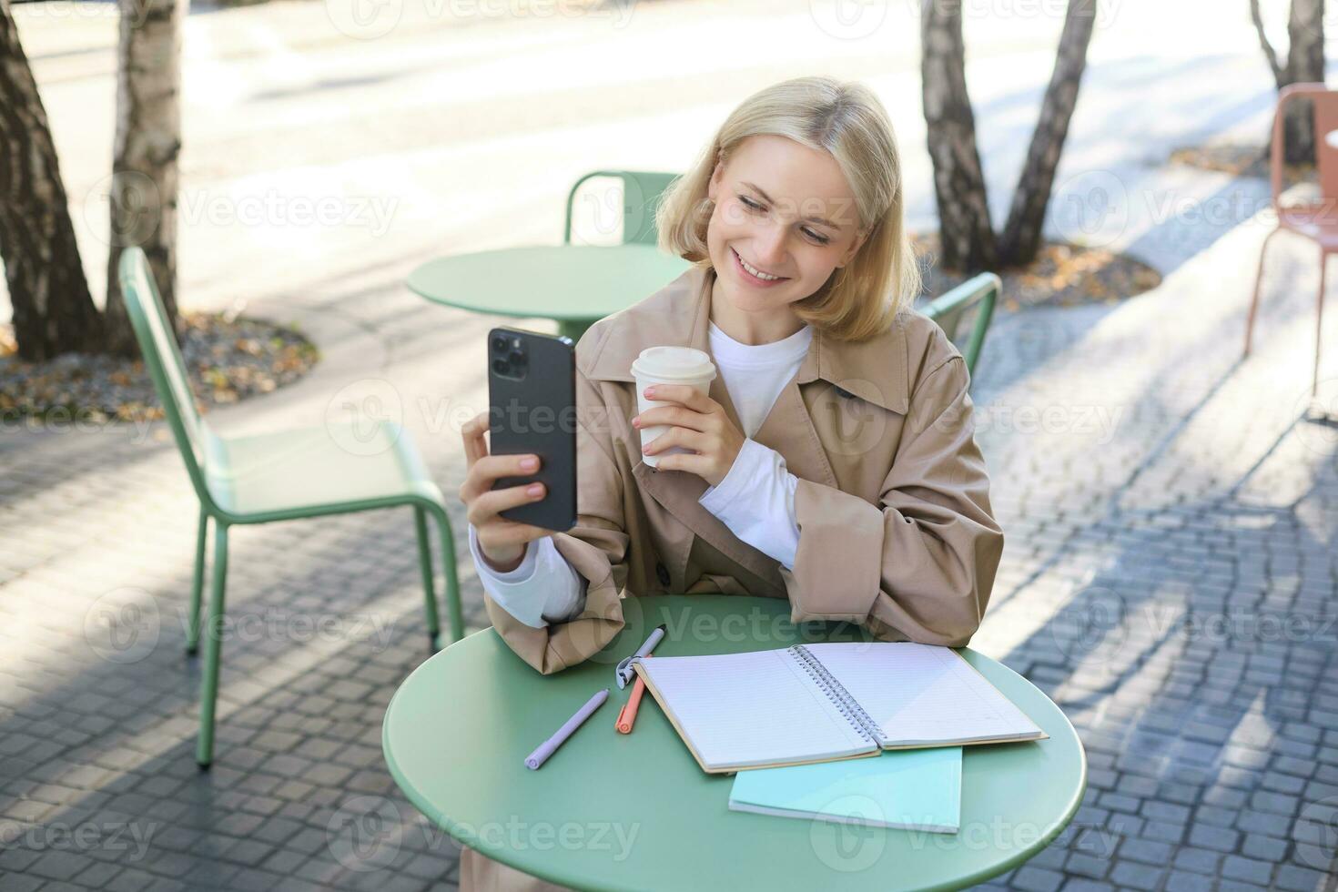 Image of stylish young woman, student taking selfie in cafe on street, posing with her favourite drink, enjoying coffee and making content for social media blog photo