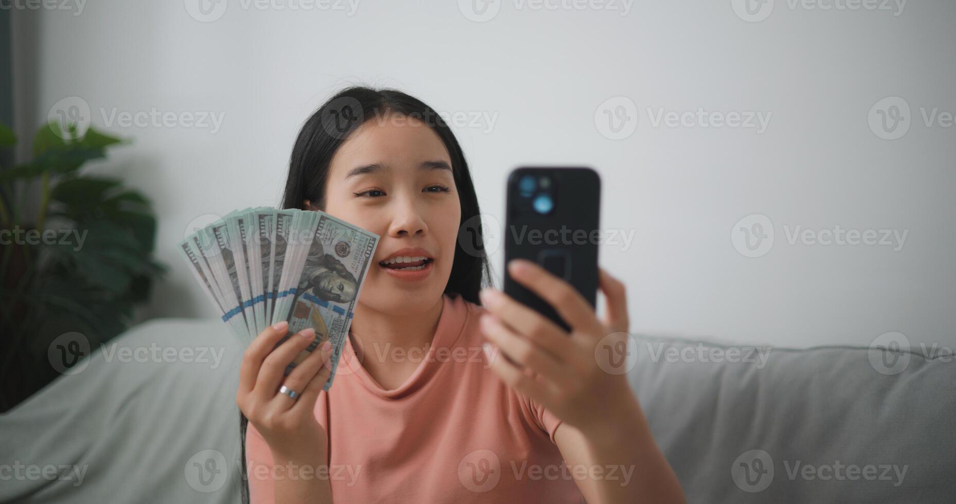 Portrait of Happy young asian woman sitting in an excited state, holding an online smartphone and dollar bills in her hand, saves money photo