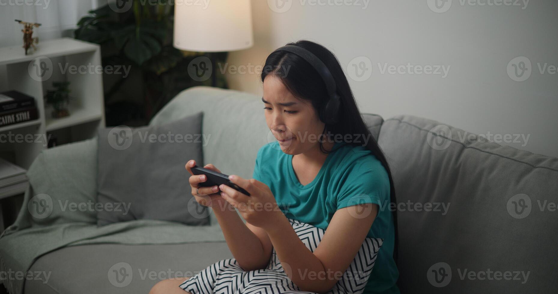 Portrait of Young Asian woman wearing glasses and headphones enjoys playing online esport games on smartphone sitting on sofa in the living room at home,Gamer lifestyle concept. photo