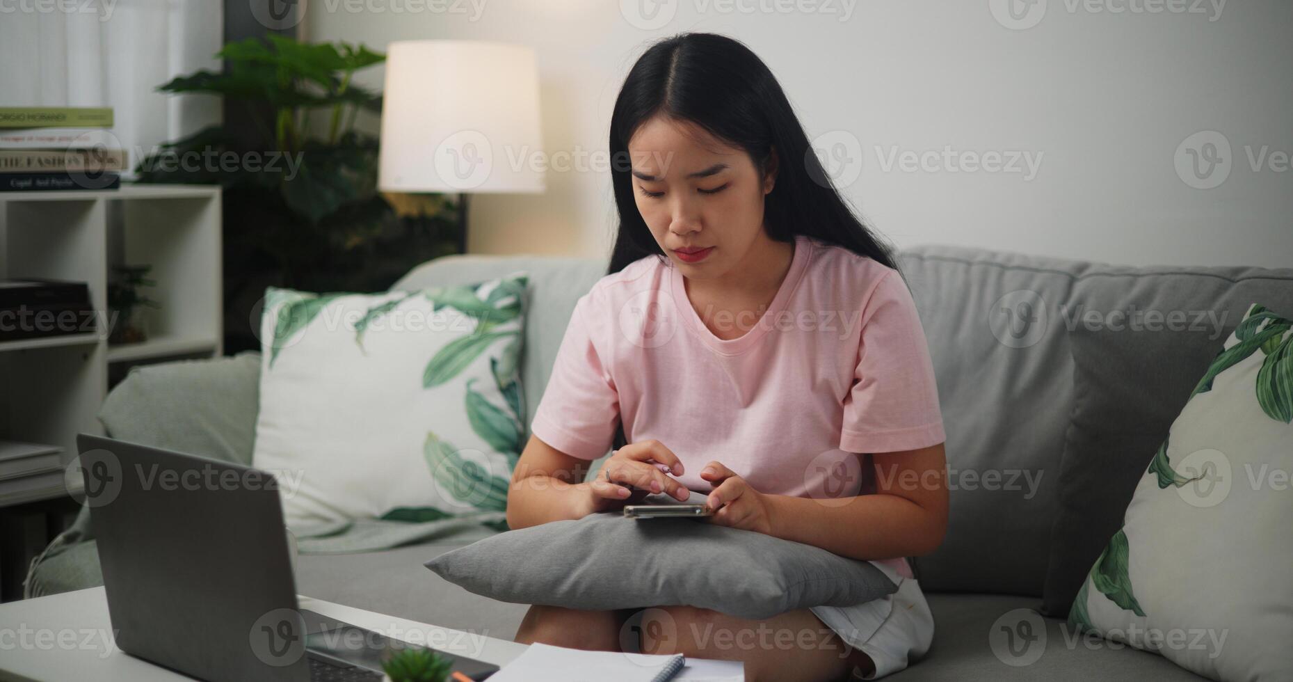 Portrait of Young Asian woman checks her finances using a calculator on smartphone and makes notes in a notebook while sitting on the sofa at home,Home finance photo