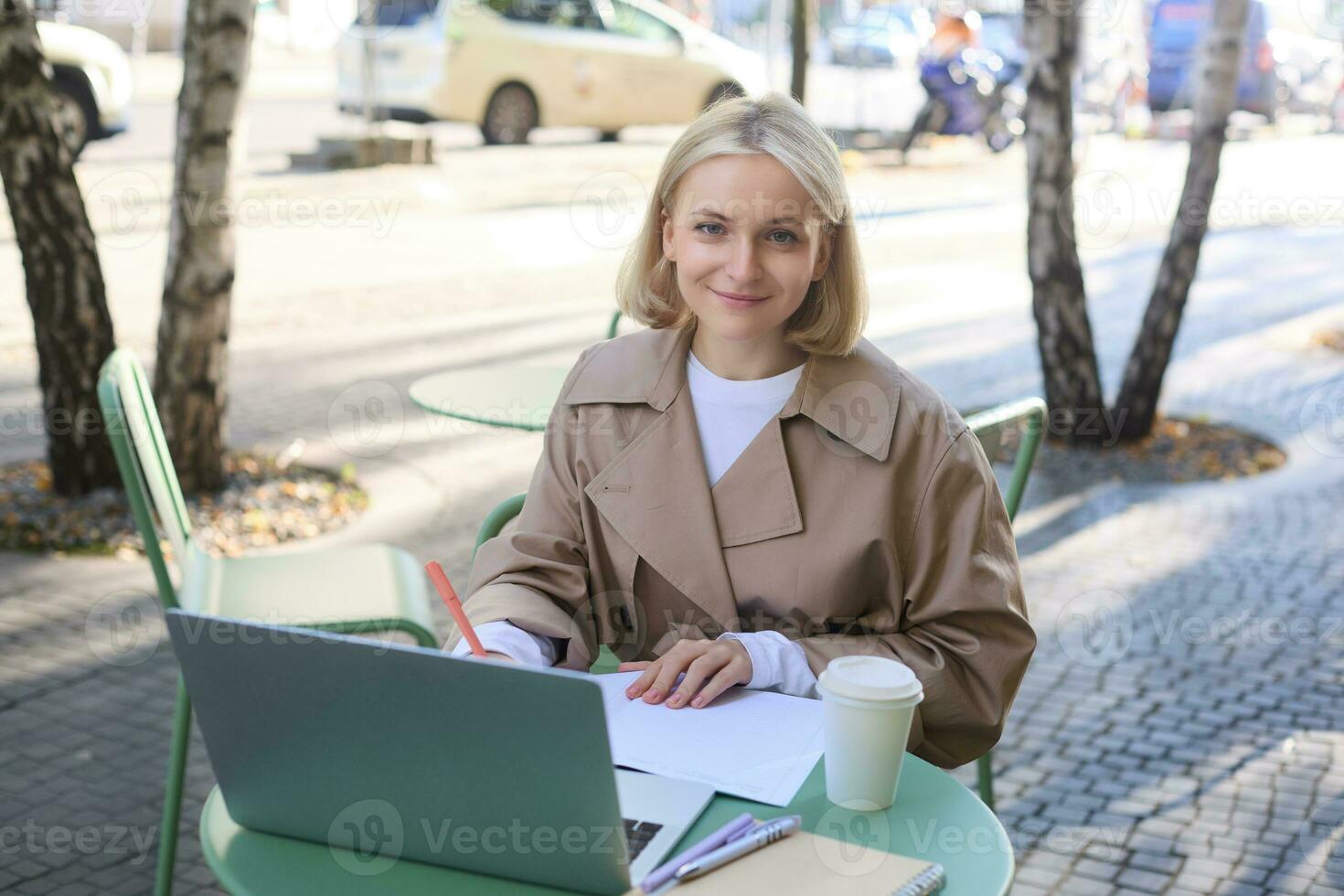 Image of young woman studying outdoors, sitting in cafe, making notes while listening to online lecture using laptop, making notes in notebook, writing photo
