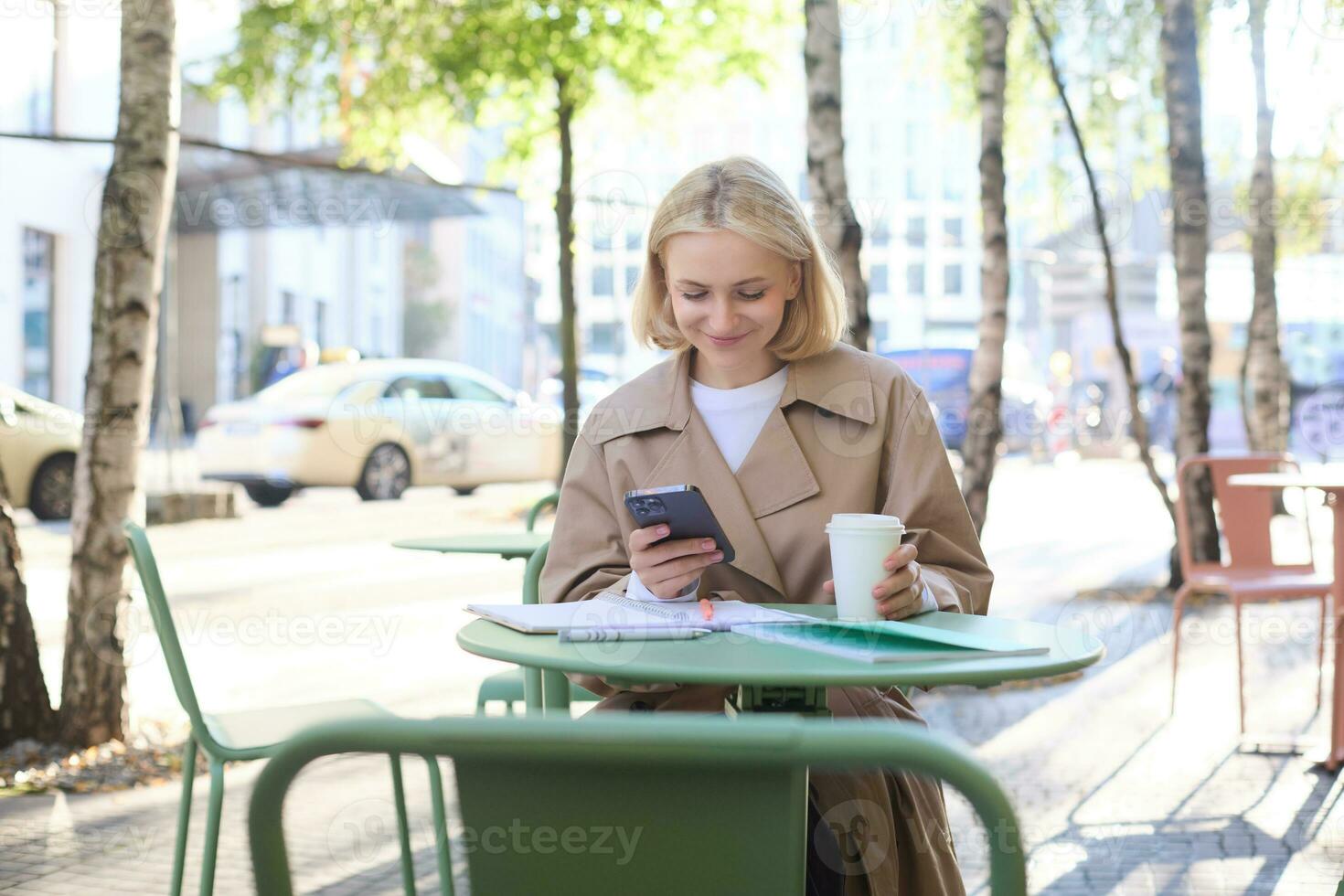 Image of young woman drinking coffee, sitting in outdoor cafe with smartphone, sending message, reading notification on mobile phone and smiling photo