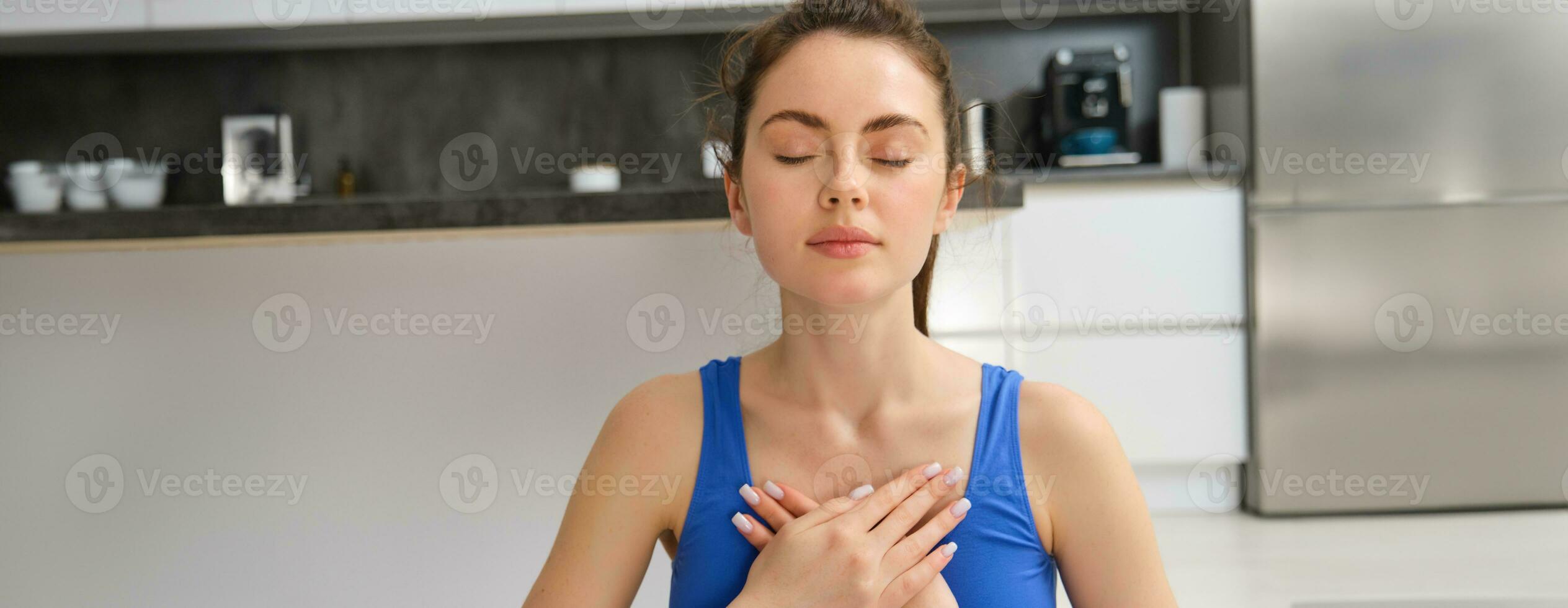 Image of calm and relaxed woman meditating, doing breathing practices, holding hands on chest during yoga session at home photo