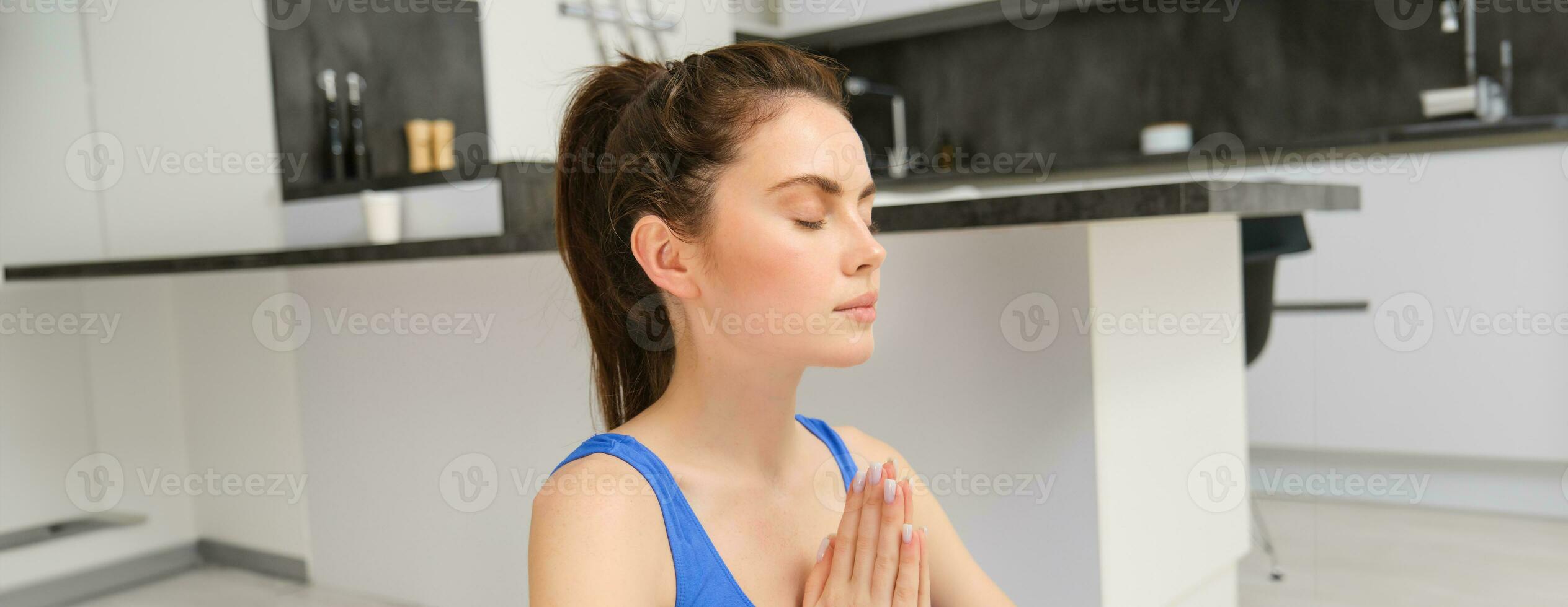 Sport and recreation. Close up portrait of beautiful girl does yoga at home, clasps her hands and meditates, sits on rubber mat in kitchen, makes asana, relaxing photo