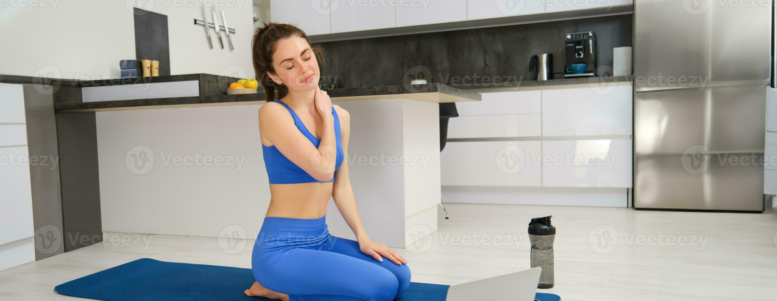 Portrait of fit and healthy woman doing fitness exercises, online yoga class from home, sitting with laptop on sports mat and massaging her neck photo