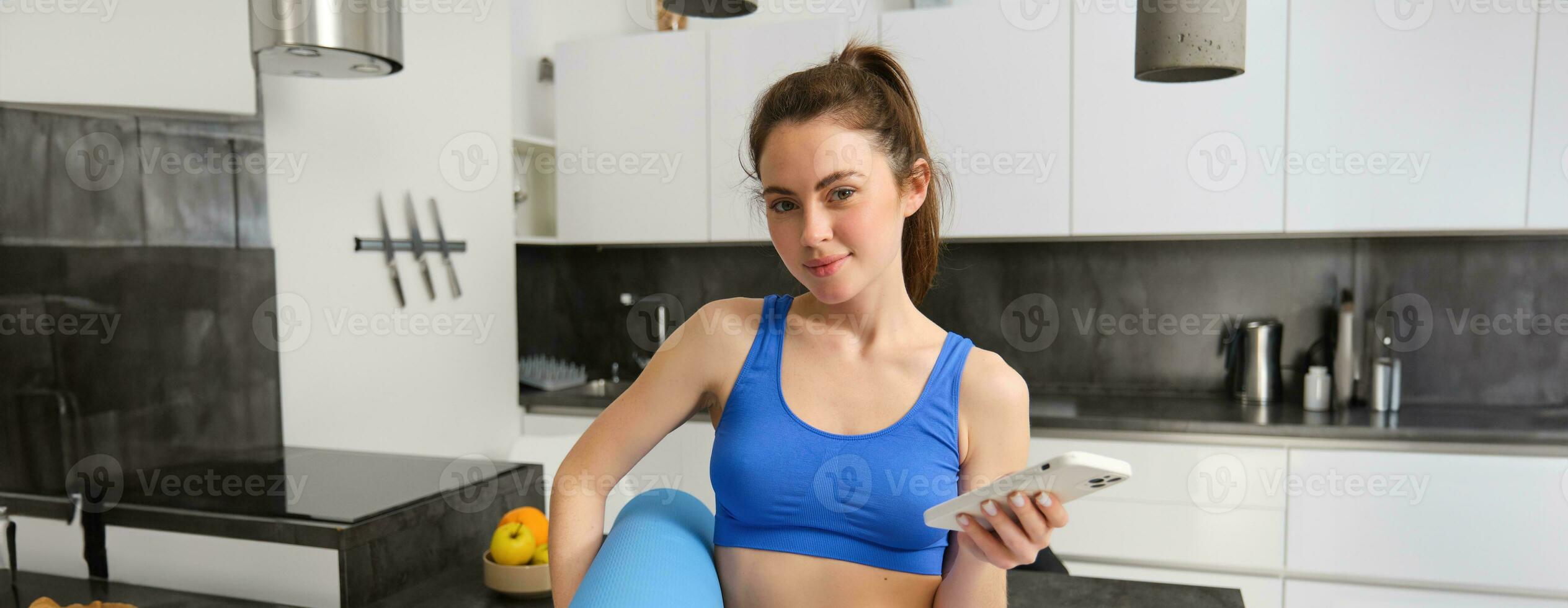 Sportwoman workout in her living room, holds rubber mat and smartphone, using mobile sports app to do fitness exercises at home photo