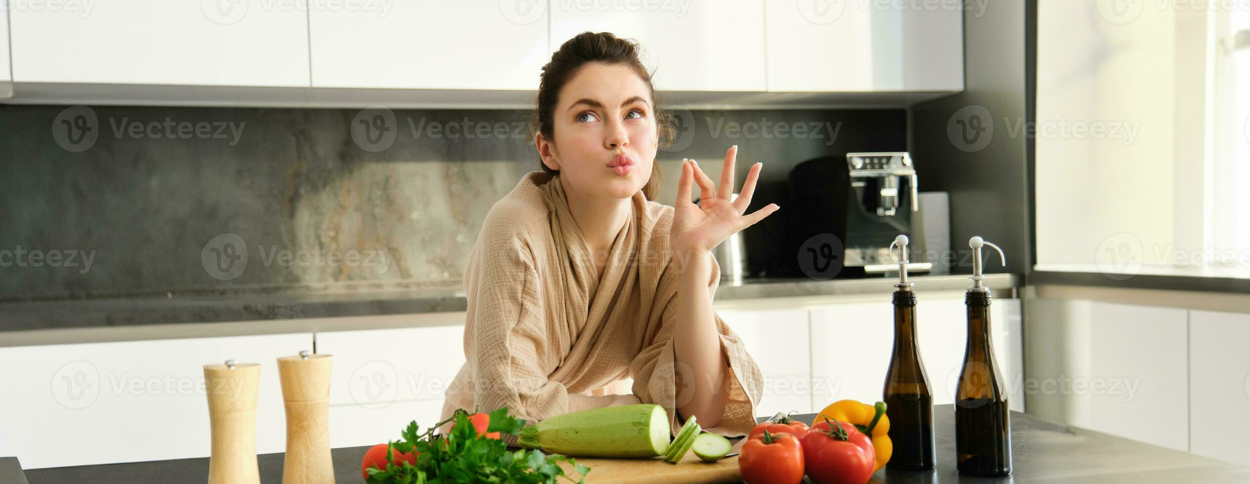 retrato de hermosa joven mujer en bata de baño, Cocinando comida para familia, muestra cocineros beso, bueno firmar, haciendo alimento, preparando vegetariano cena, el cortar vegetales foto