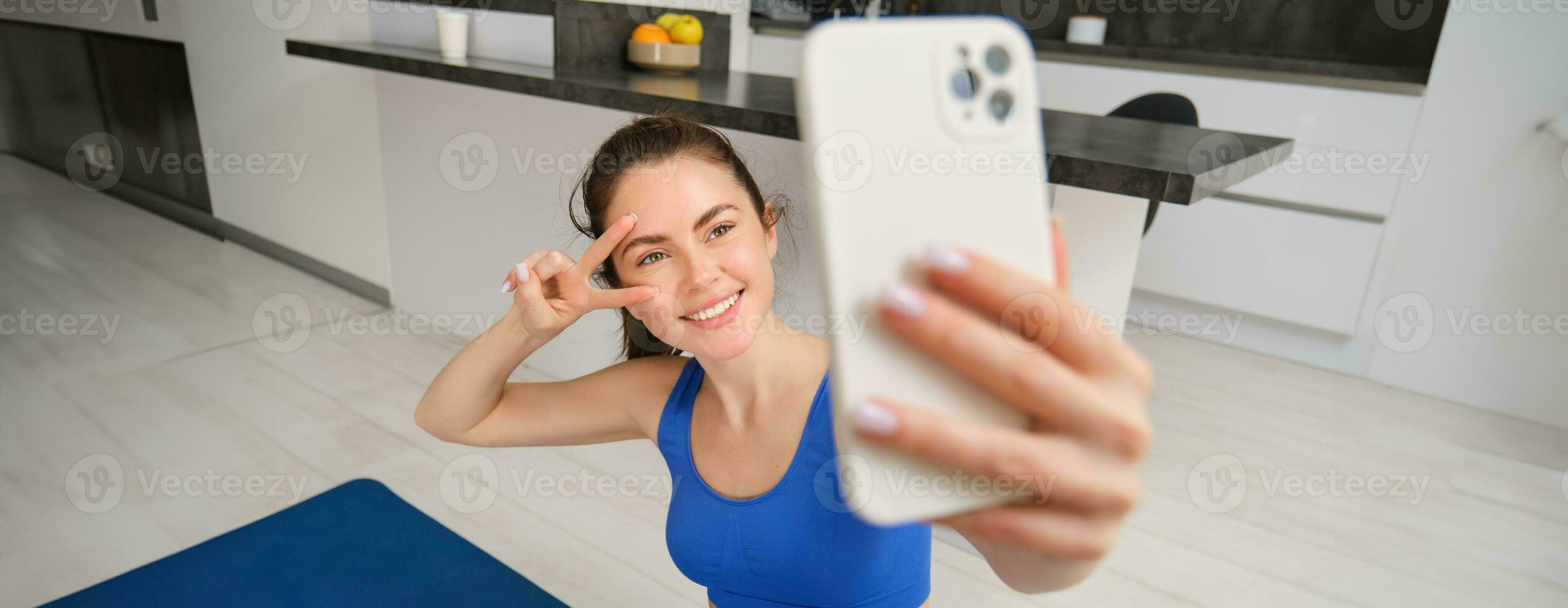 Portrait of girl takes selfie on smartphone while does her workout training. Young woman does her exercises at home, takes selfie for sports blog, sits in living room photo