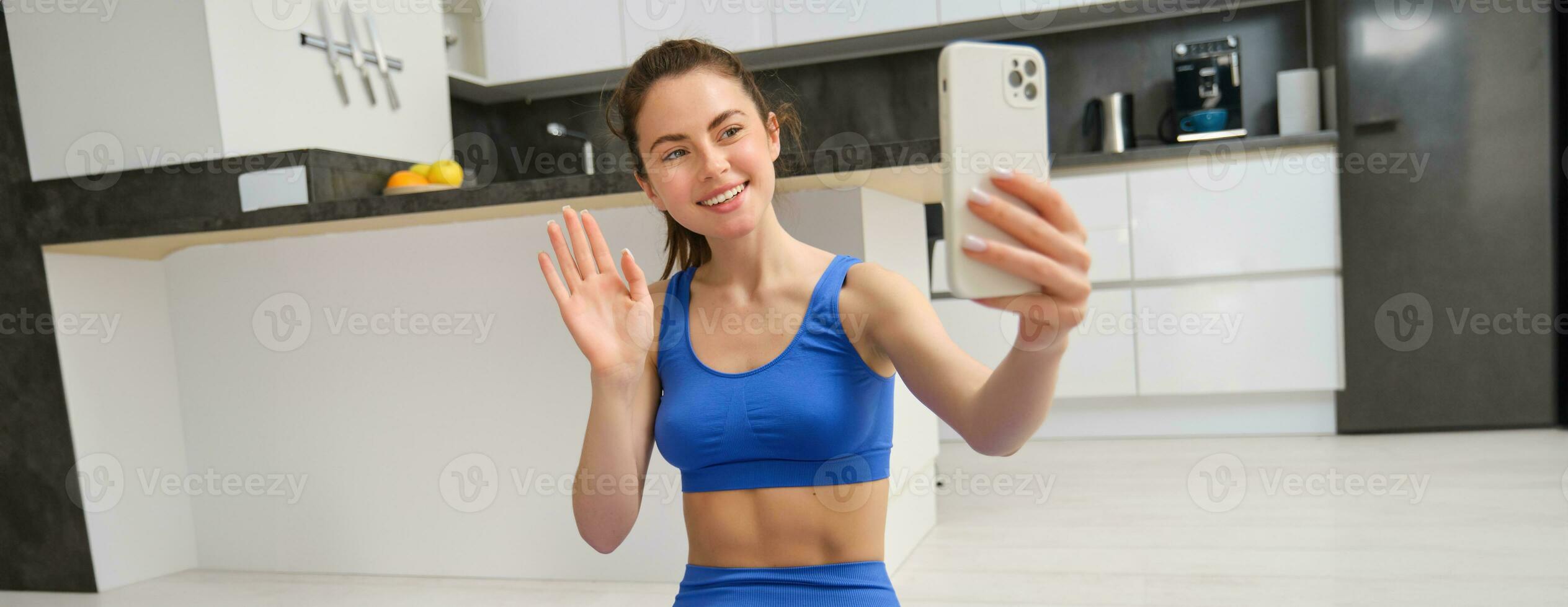 Smiling beautiful girl records video while doing sports at home, looks at smartphone, takes selfie on mobile phone, workout indoors in blue leggings and sportsbra photo