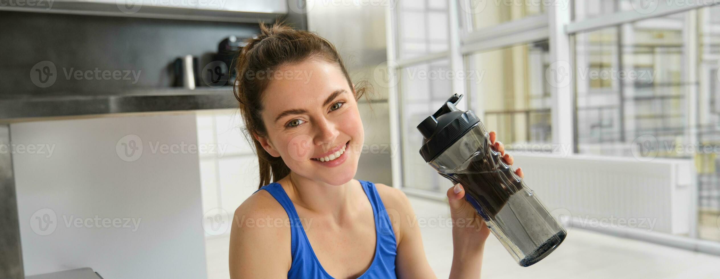 sonriente aptitud niña bebidas agua en el medio de formación sesión, sonriente y mirando feliz, rutina de ejercicio desde hogar en azul sujetador deportivo foto