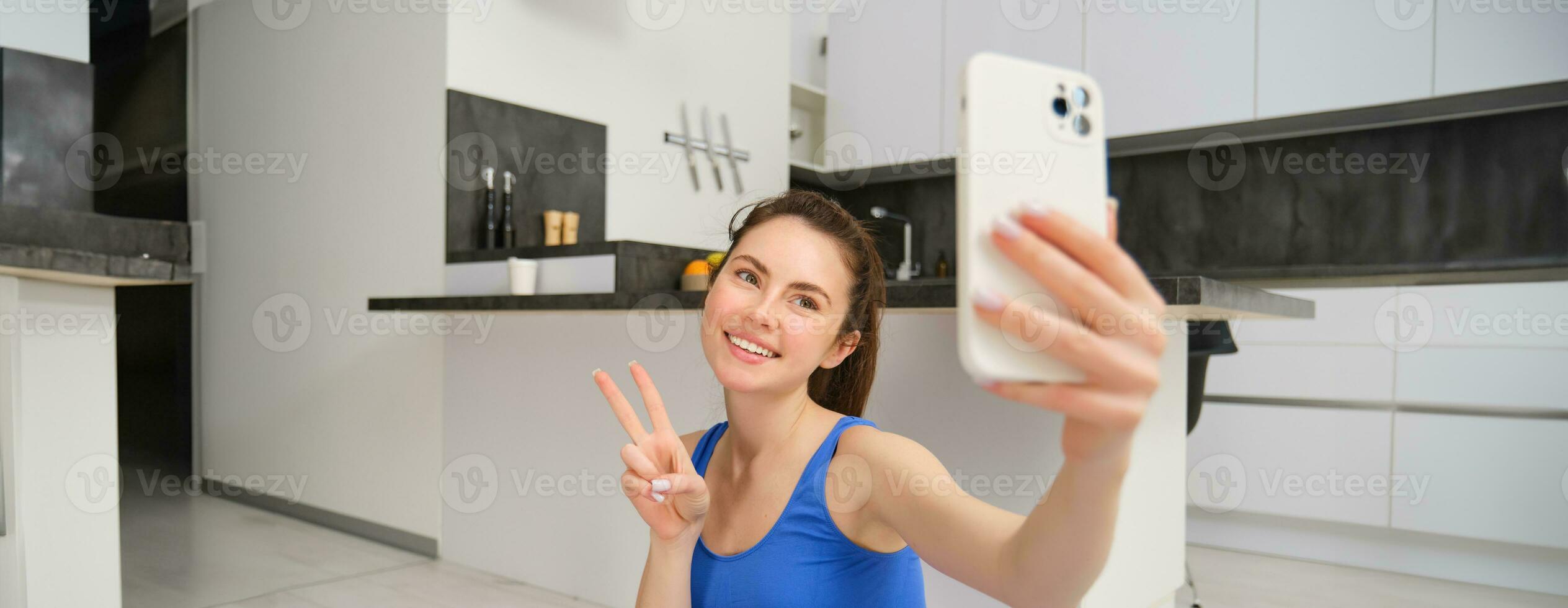 Close up portrait of beautiful brunette sportswoman, taking selfie on smartphone, posing on fitness mat with mobile phone, records her fitness exercises, workout training photo