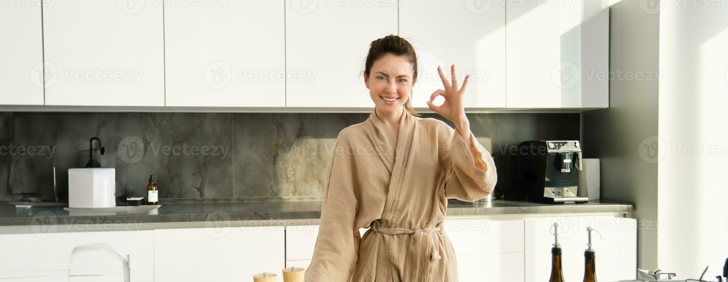 Portrait of smiling brunette woman cooking healthy dinner, showing okay sign, wife standing in bathrobe in kitchen, preparing food, chopping vegetables on board photo