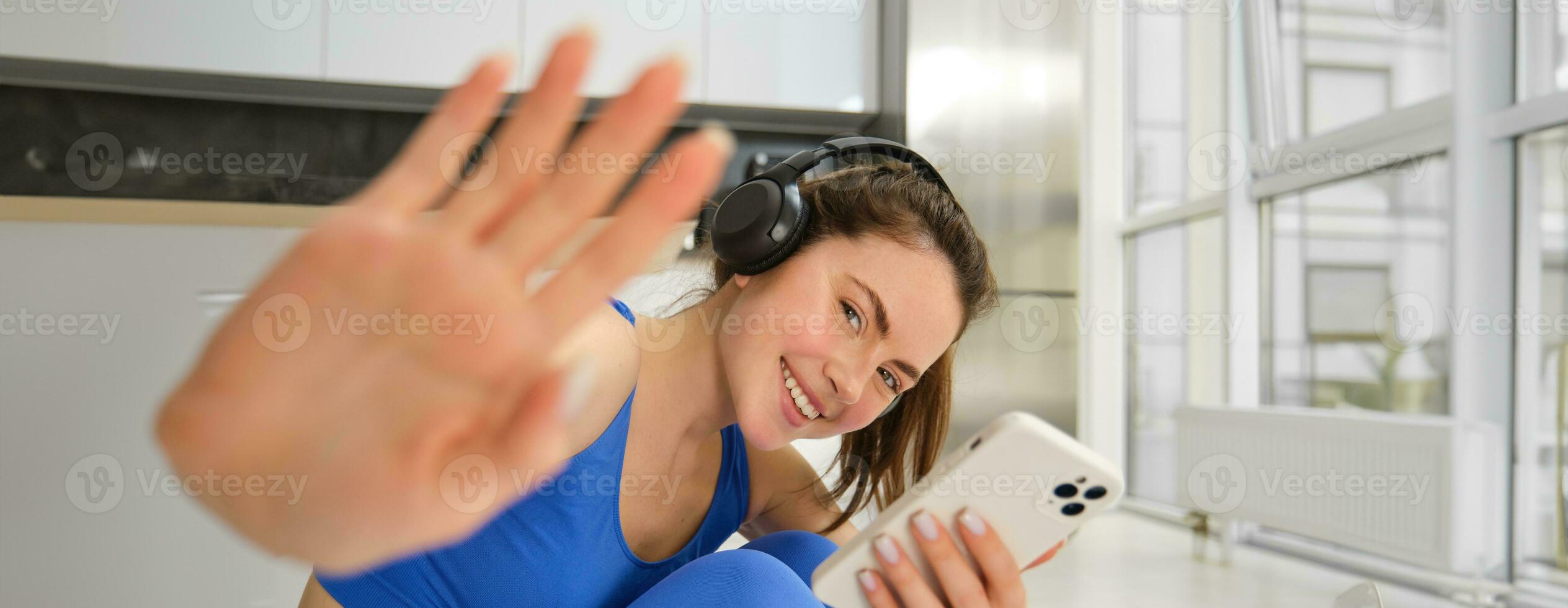 Portrait of beautiful brunette girl, hides from camera, puts her hand towards camera, listens music in headphones and holds smartphone, watches exercses online on mobile phone, smiles photo