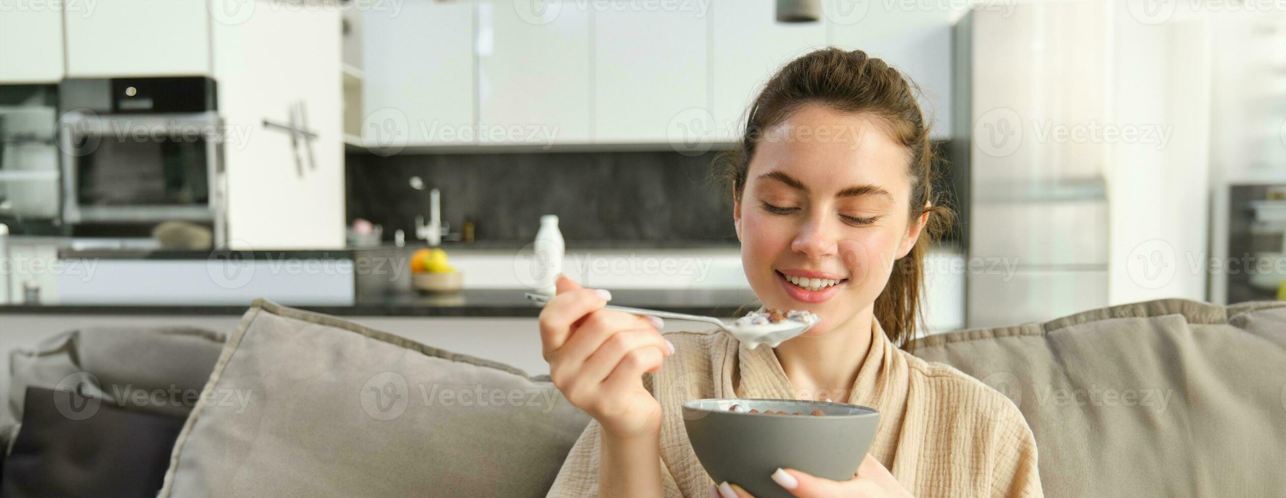 Image of smiling, happy young woman eating breakfast, holding bowl of cereals with milk, having meal at home photo