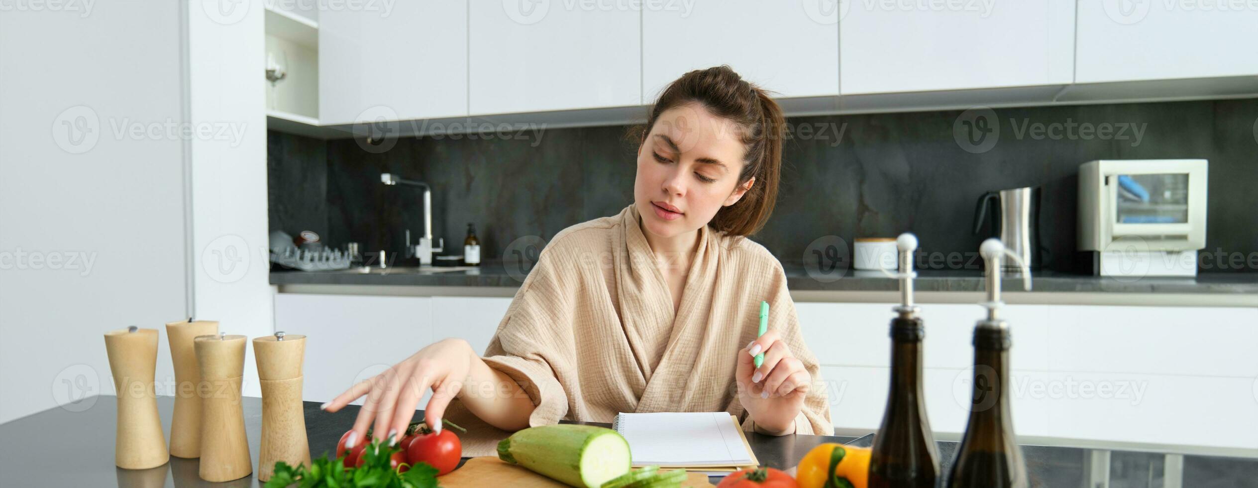 Portrait of woman cooking in the kitchen, sitting in front of vegetables, tomatoes zucchini and parsley, making list of groceries, writing down recipe, wearing bathrobe photo