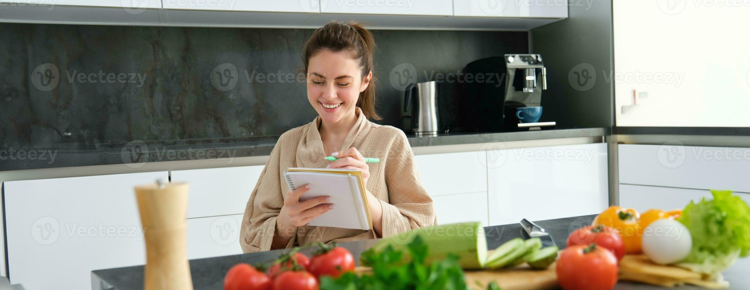 Portrait of beautiful, smiling young woman making list of meals, writing down recipe, sitting in the kitchen with vegetables, doing house errands photo
