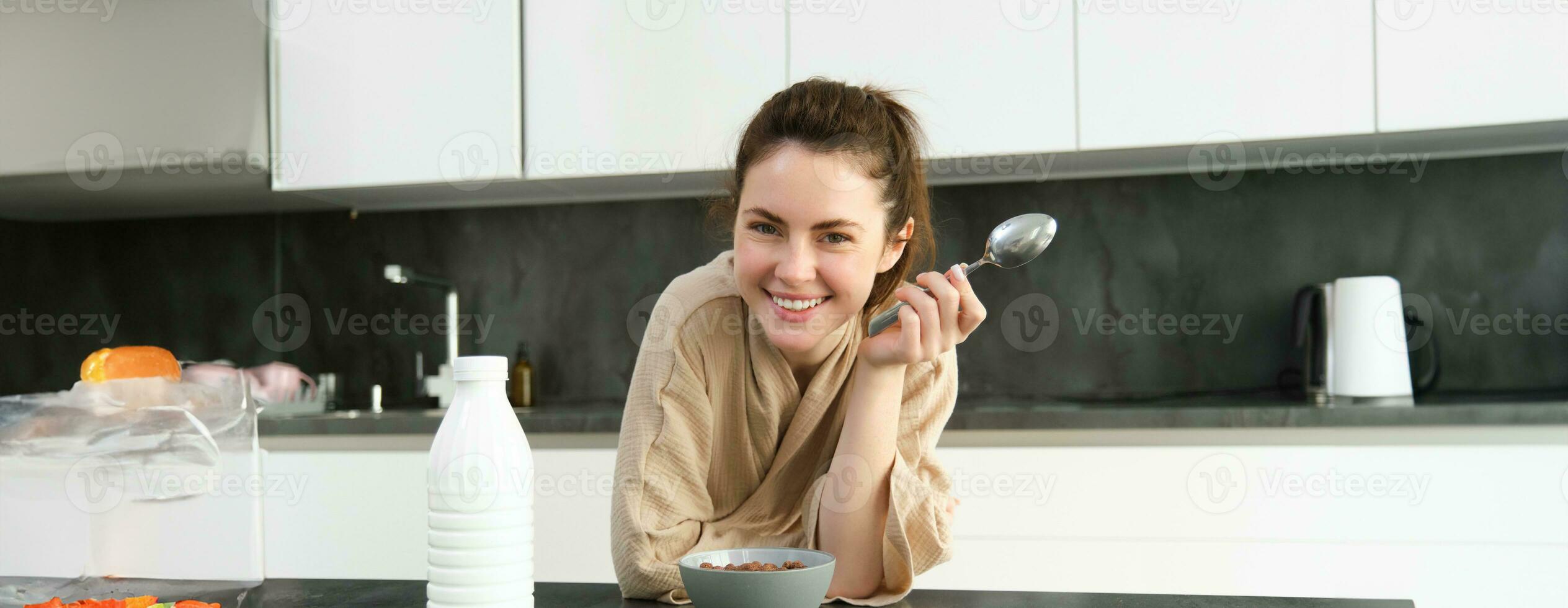 Laughing, beautiful woman in bathrobe, eats cereals with milk, holding spoon, smiling and looking happy, posing in kitchen and leaning on worktop photo