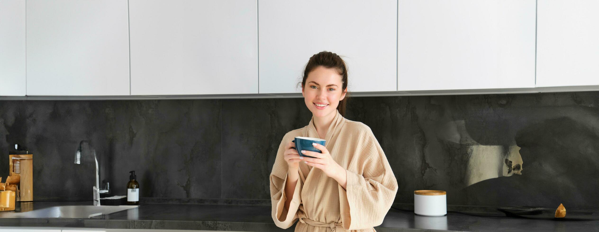 retrato de hermosa ama de casa, joven mujer en bata de baño, participación taza de café, Bebiendo té en el cocina, disfrutando Mañana foto