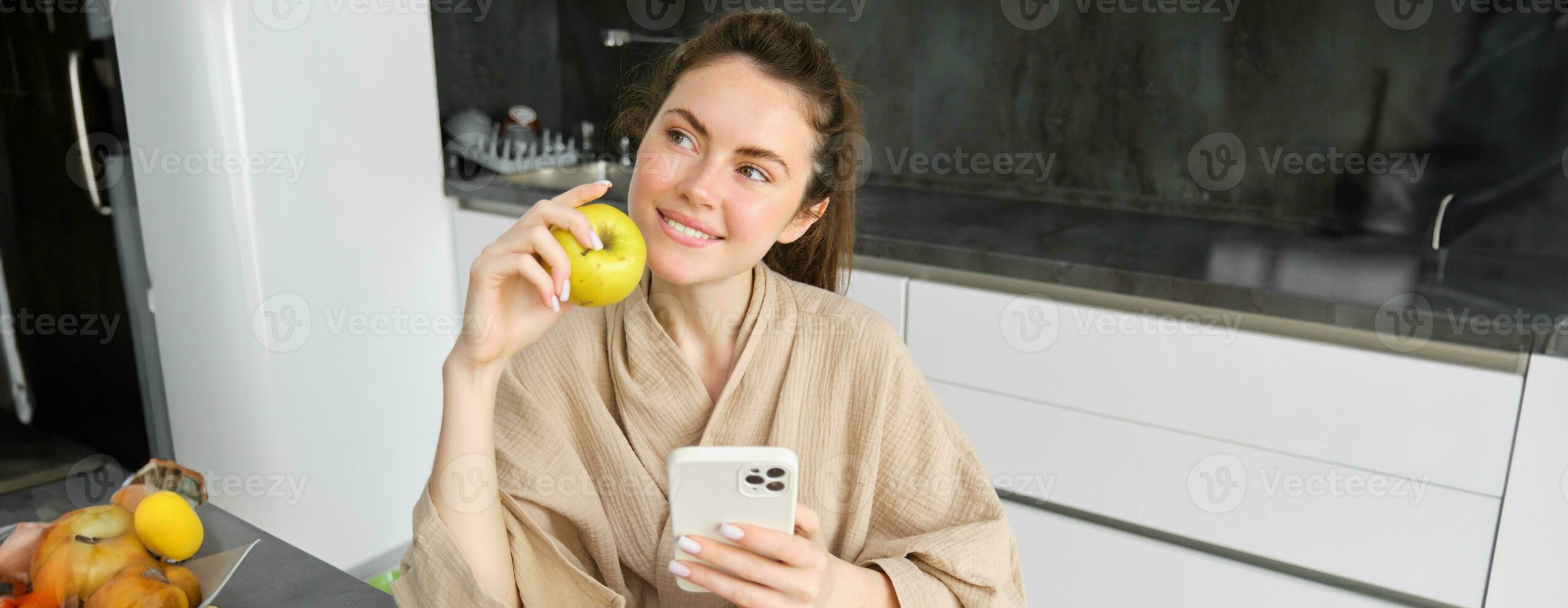 Close up portrait of happy young woman in bathrobe, sitting in the kitchen and using mobile phone, holding an apple, order fruits and vegetables online, using smartphone app for groceries delivery photo