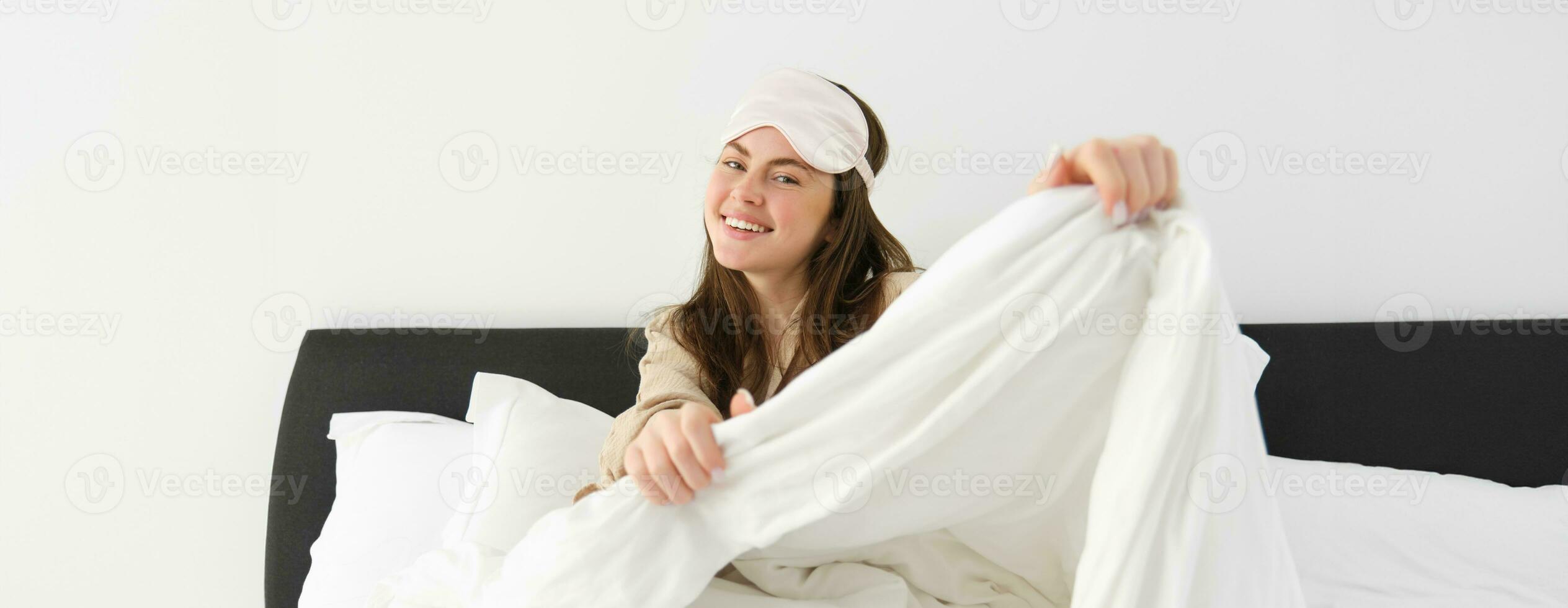 Beautiful young woman with sleeping mask on forehead, waking up in morning and smiling, resting in her bedroom, feeling lazy on weekends photo