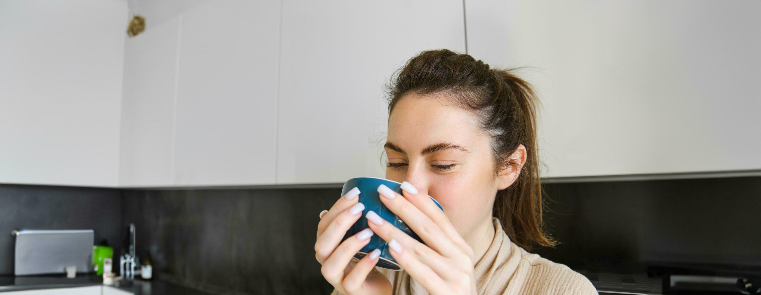 Portrait of carefree smiling woman drinking coffee, standing in kitchen with delighted, pleased face expression, waking up in the morning and having cappuccino photo