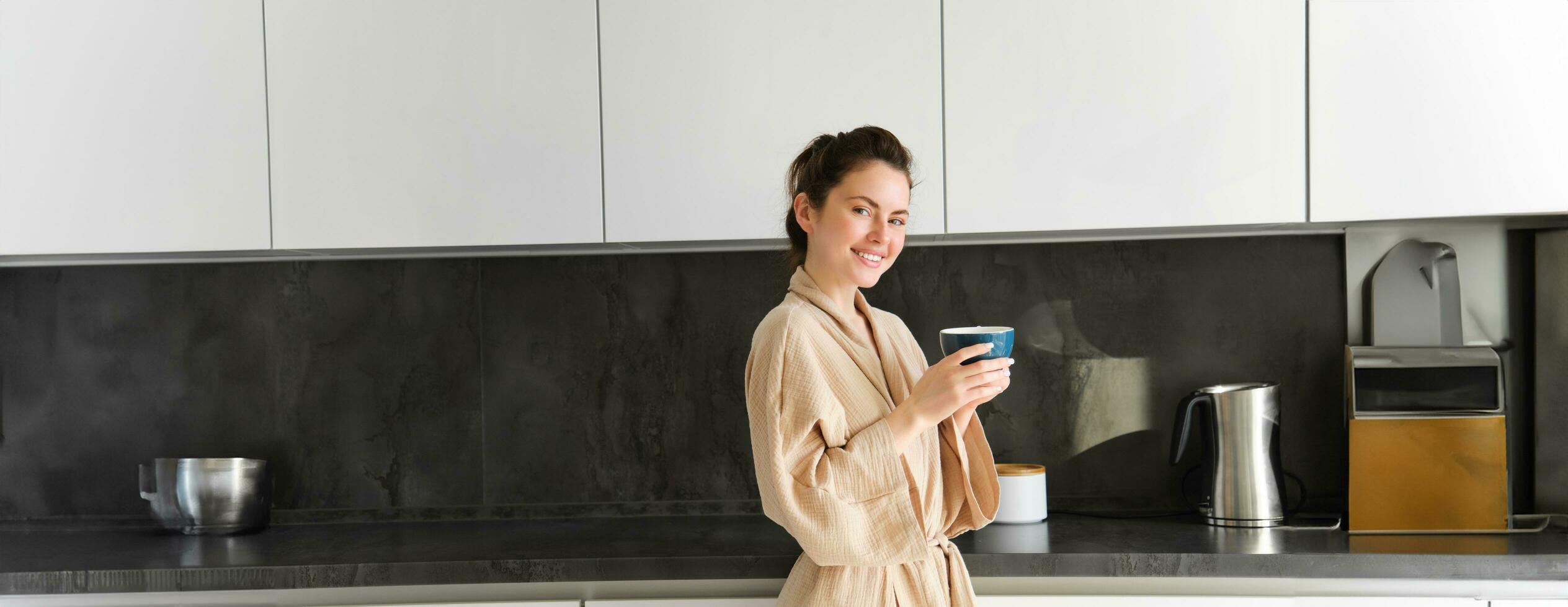Daily routine and lifestyle. Young beautiful woman in bathrobe, standing in kitchen with cup of coffee, drinking tea, smiling and looking happy photo