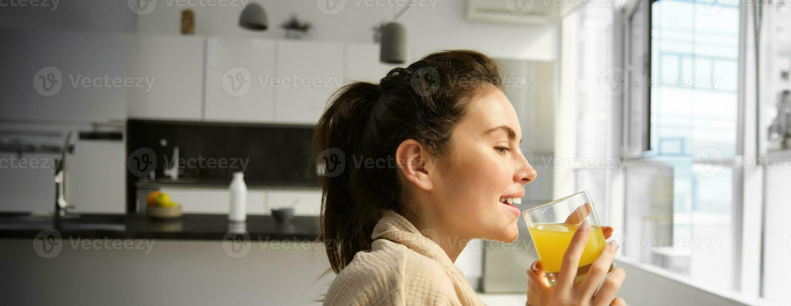 Close up portrait of happy young woman, enjoys relaxing day at home, drinks glass of orange juice and smiles, looks outside window photo