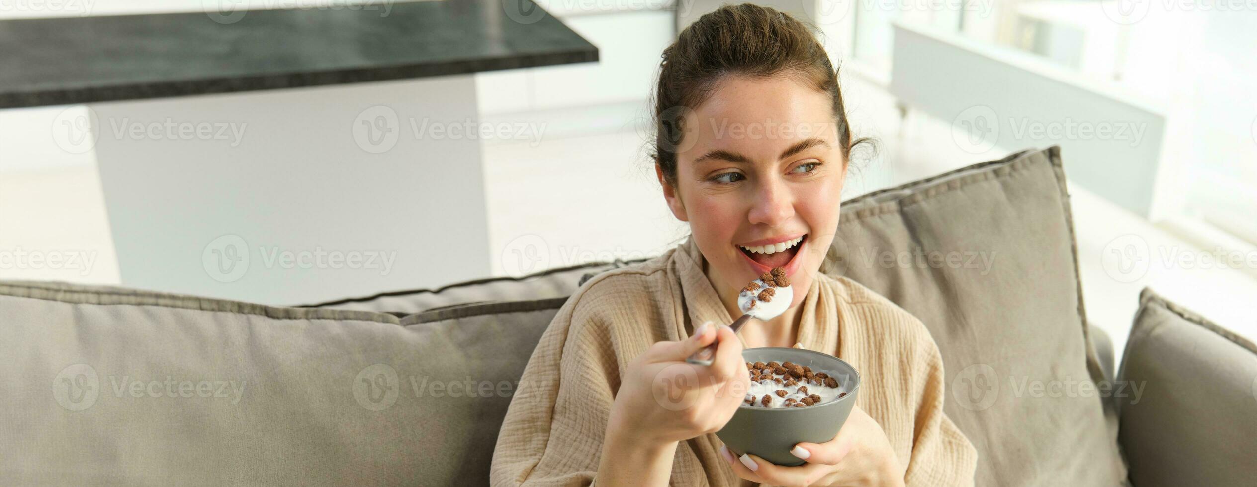 Portrait of woman on sofa in bathrobe, eating cereals with milk, watching tv and having breakfast photo