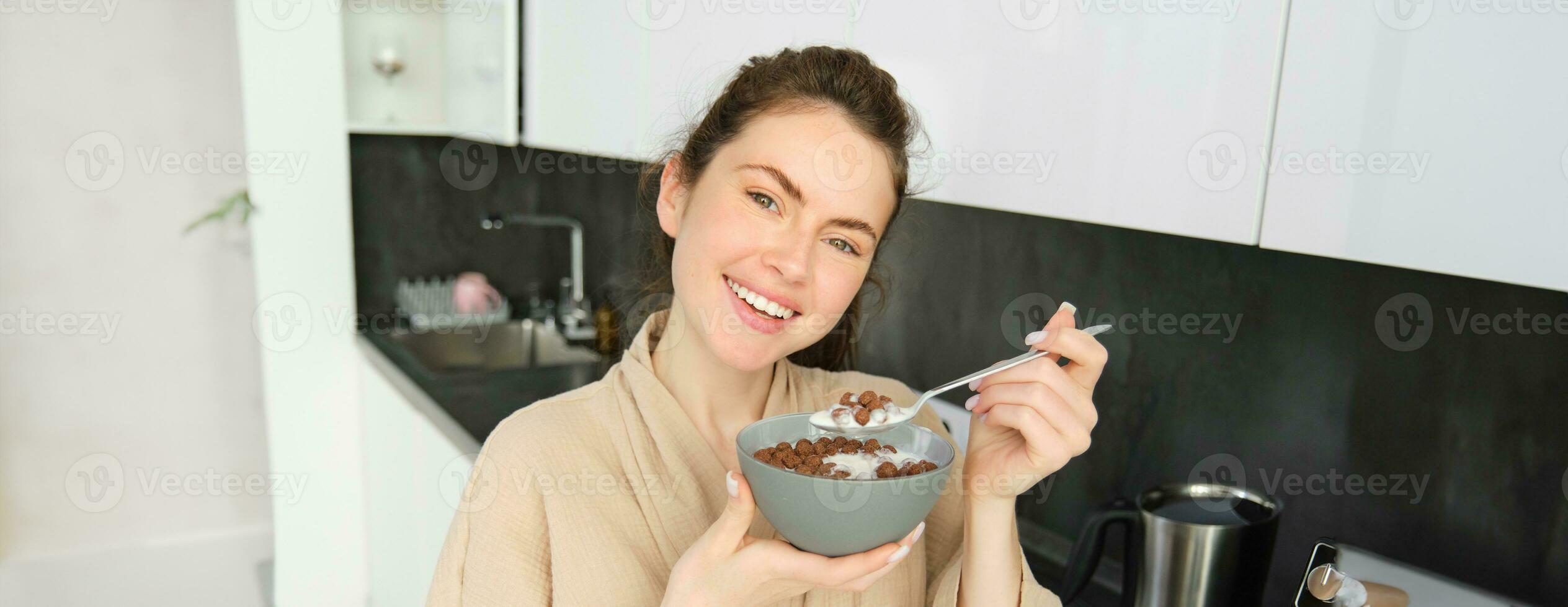 Close up of beautiful smiling woman in bathrobe, standing in kitchen near worktop, eating bowl of chocolate cereals with milk, holding spoon and looking happy at camera photo