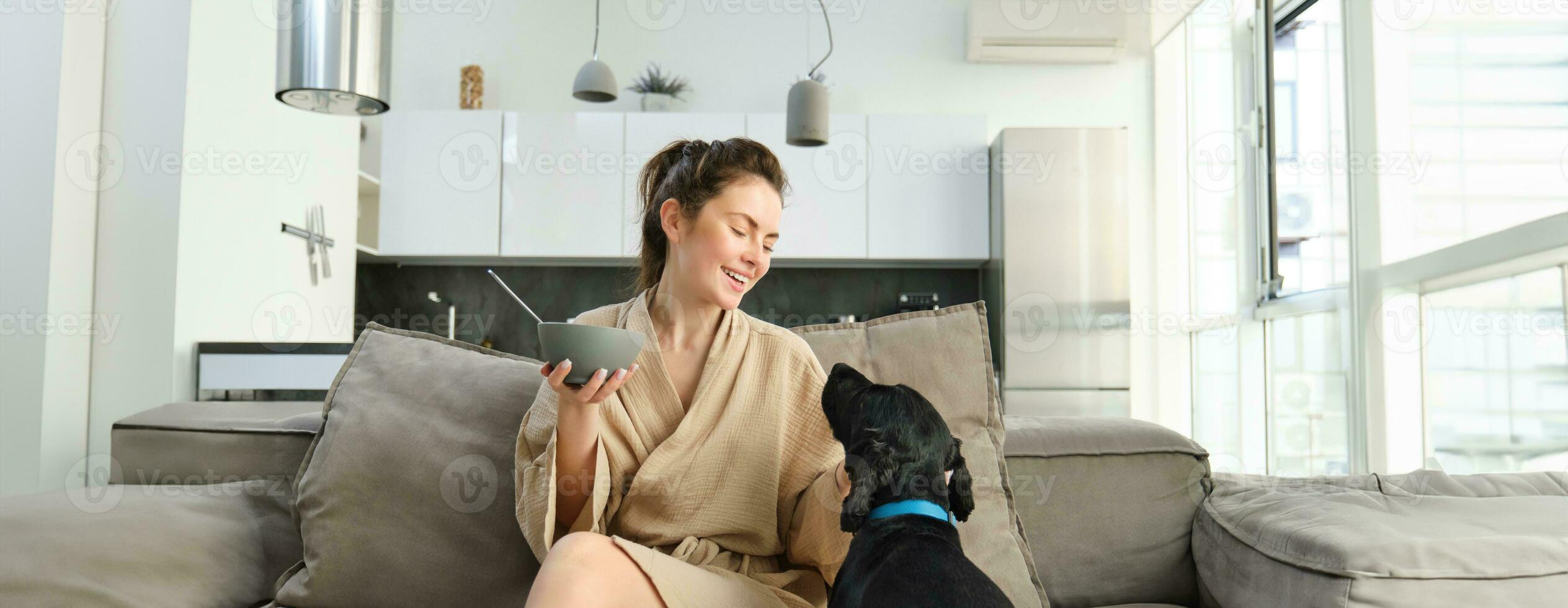 acogedor mañanas con mejor amigo. joven mujer comiendo cereales para desayuno, sentado con su perro en sofá y sonriente, comenzando su Mañana con apropiado comida foto