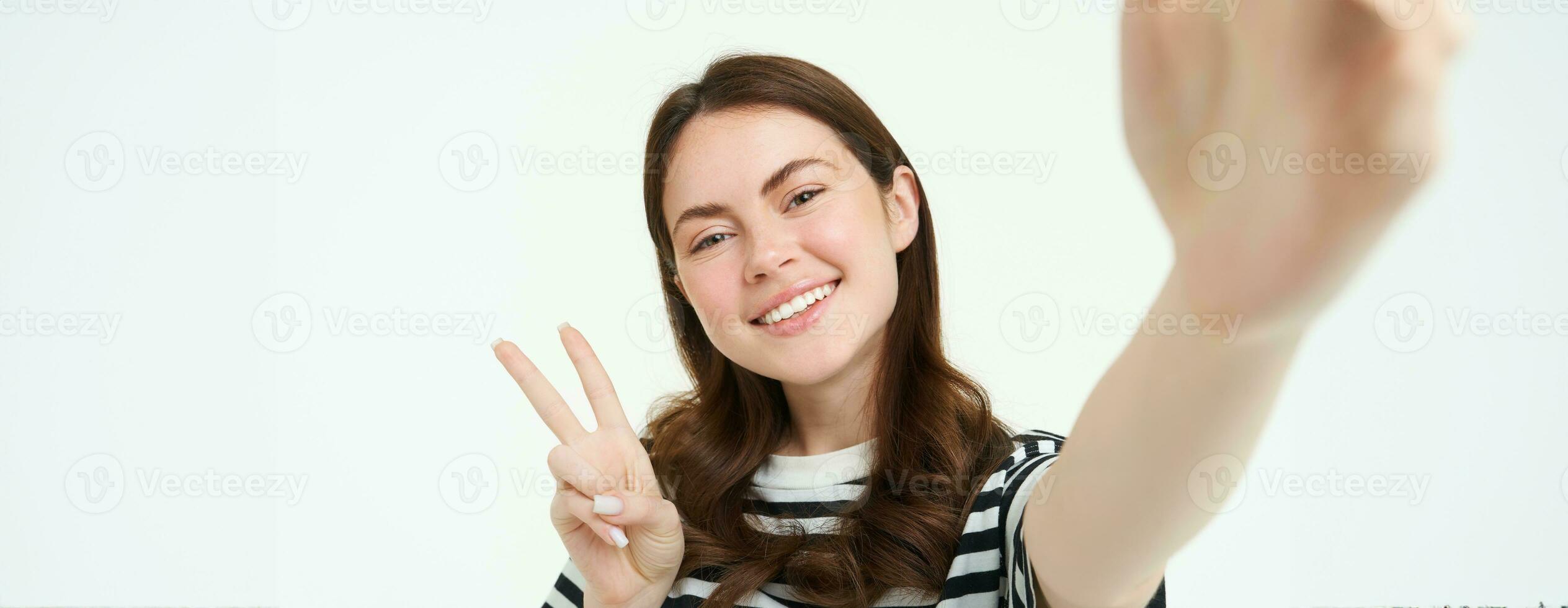 Smartphone view, woman holding mobile phone camera and taking selfie with peace, v-sign gesture, smiling at camera, posing over white background photo