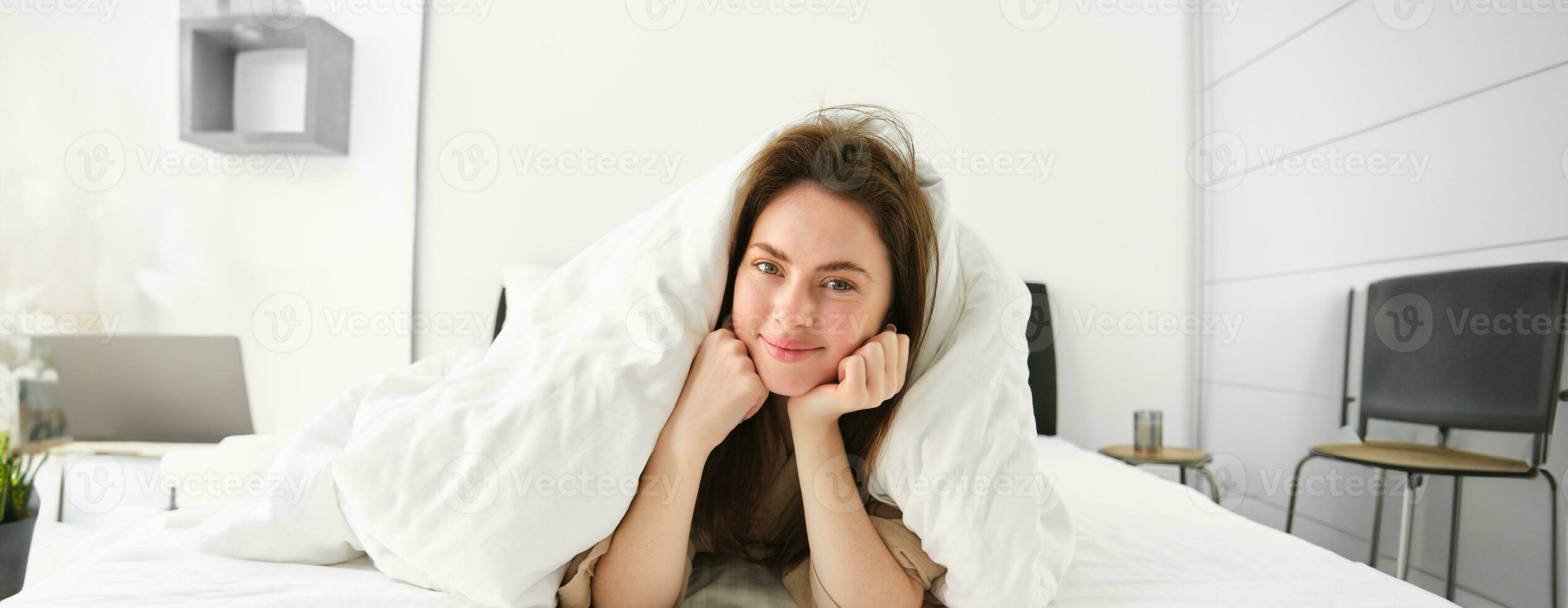 Portrait of attractive smiling woman relaxing in bed, lying covered in white sheets and warm duvet in morning, looking happy at camera photo