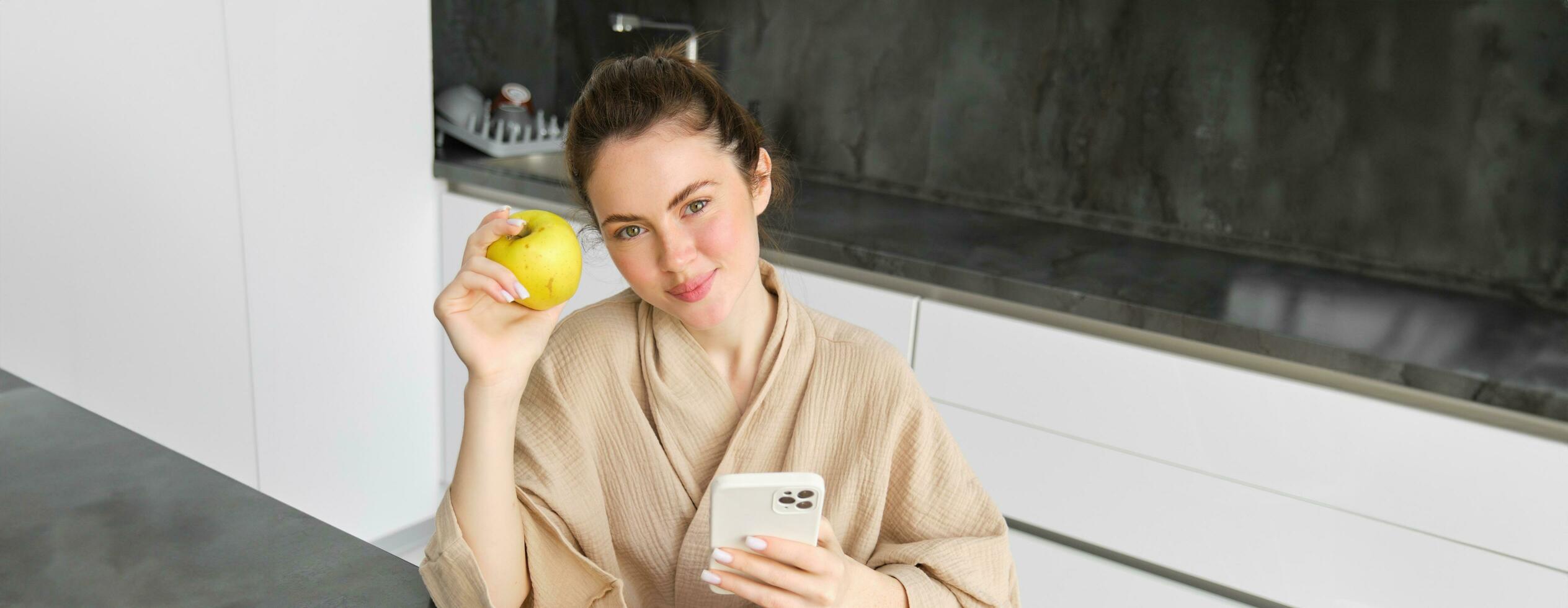 Image of attractive young woman in bathrobe, sits in kitchen, eats green apple and looks at mobile phone, uses smartphone app, orders food on application photo
