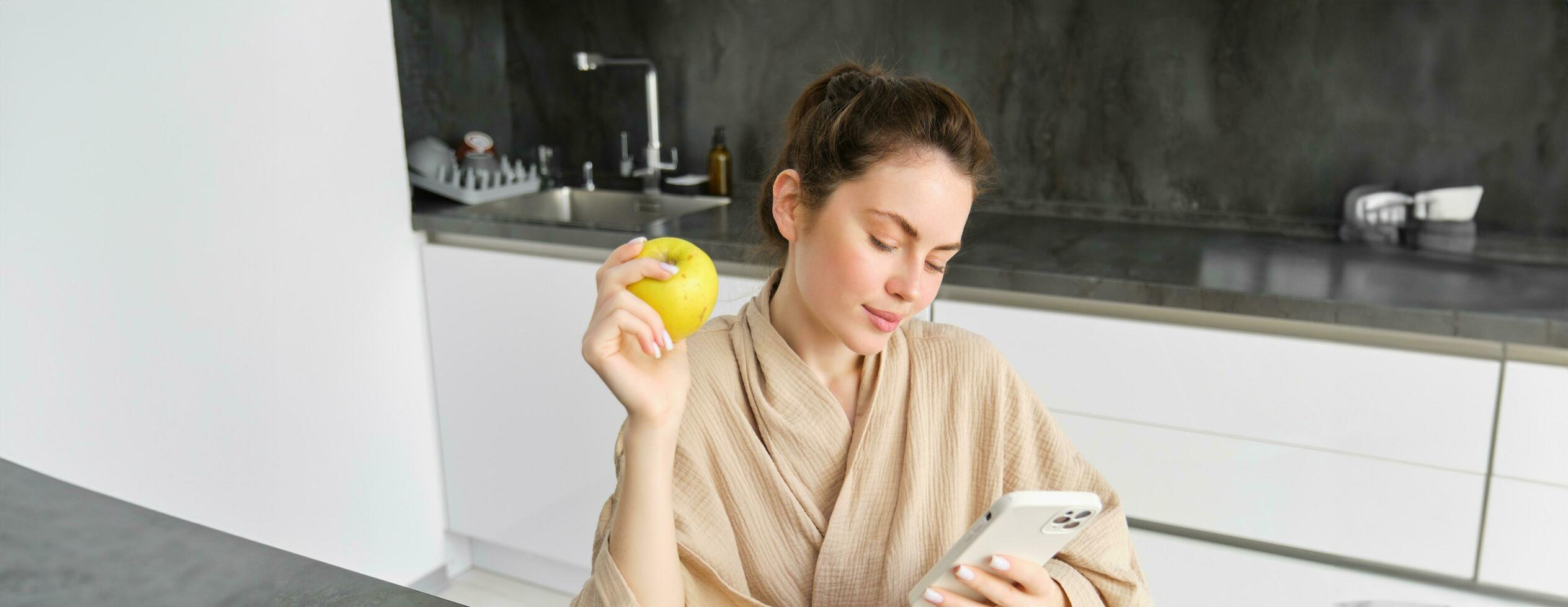 Image of attractive young woman in bathrobe, sits in kitchen, eats green apple and looks at mobile phone, uses smartphone app, orders food on application photo