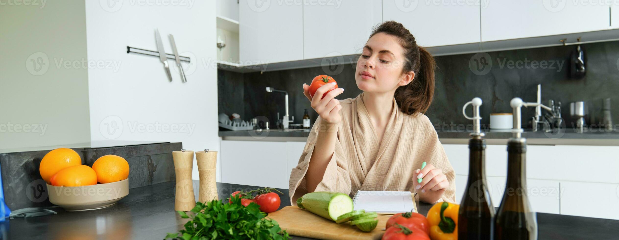 Portrait of beautiful smiling woman, writing her healthy menu, eating tomato while cooking, making grocery list, sitting in the kitchen photo