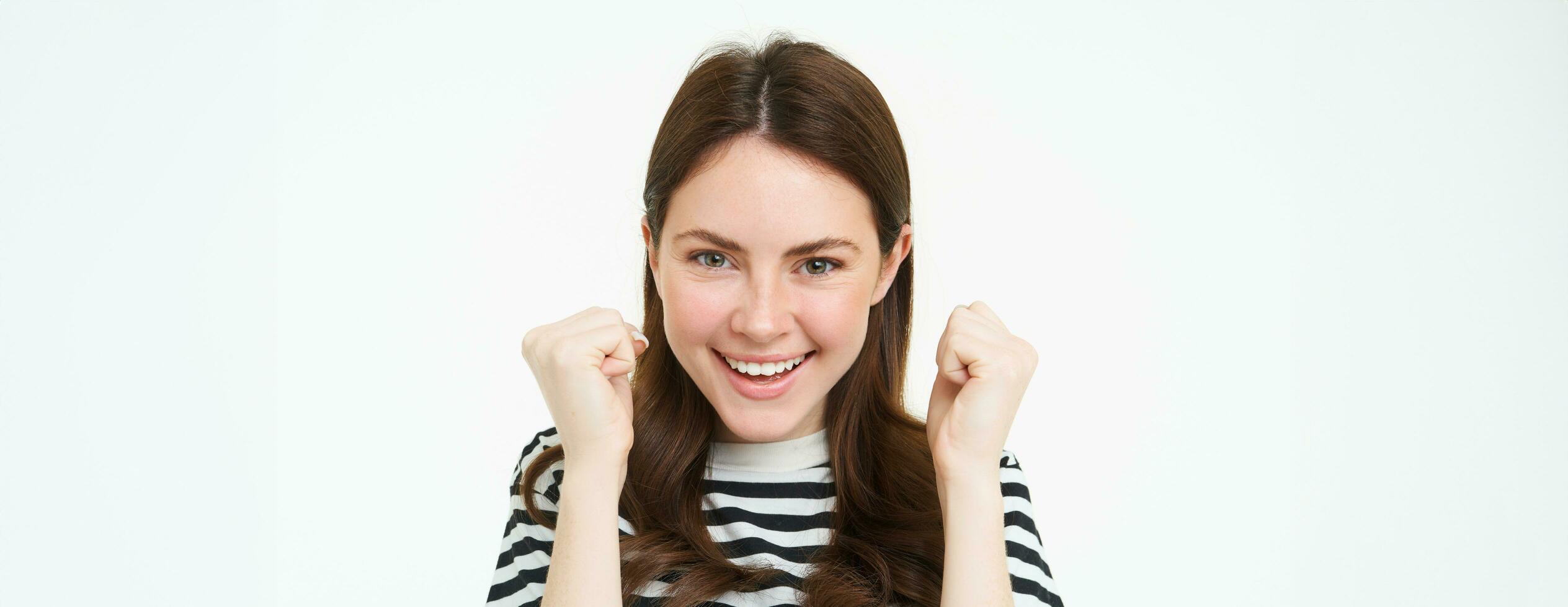 Image of young woman looking with pleased, satisfied face at camera, winning, shaking hands in triumph, celebrating victory, achievement, standing over white background photo