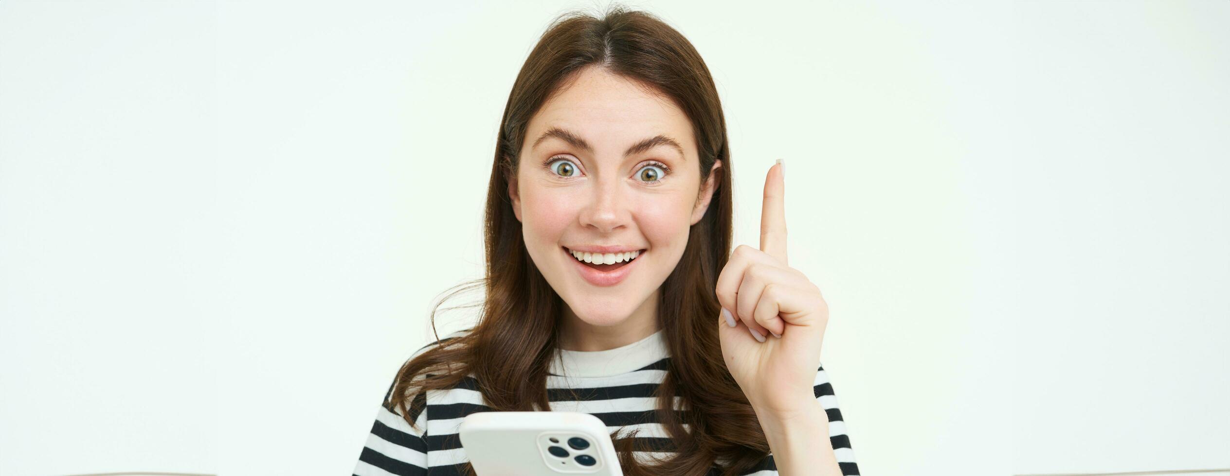 Got an idea. Smiling woman, raises her finger and holds smartphone, suggests something, makes eureka gesture, has a solution, white background photo